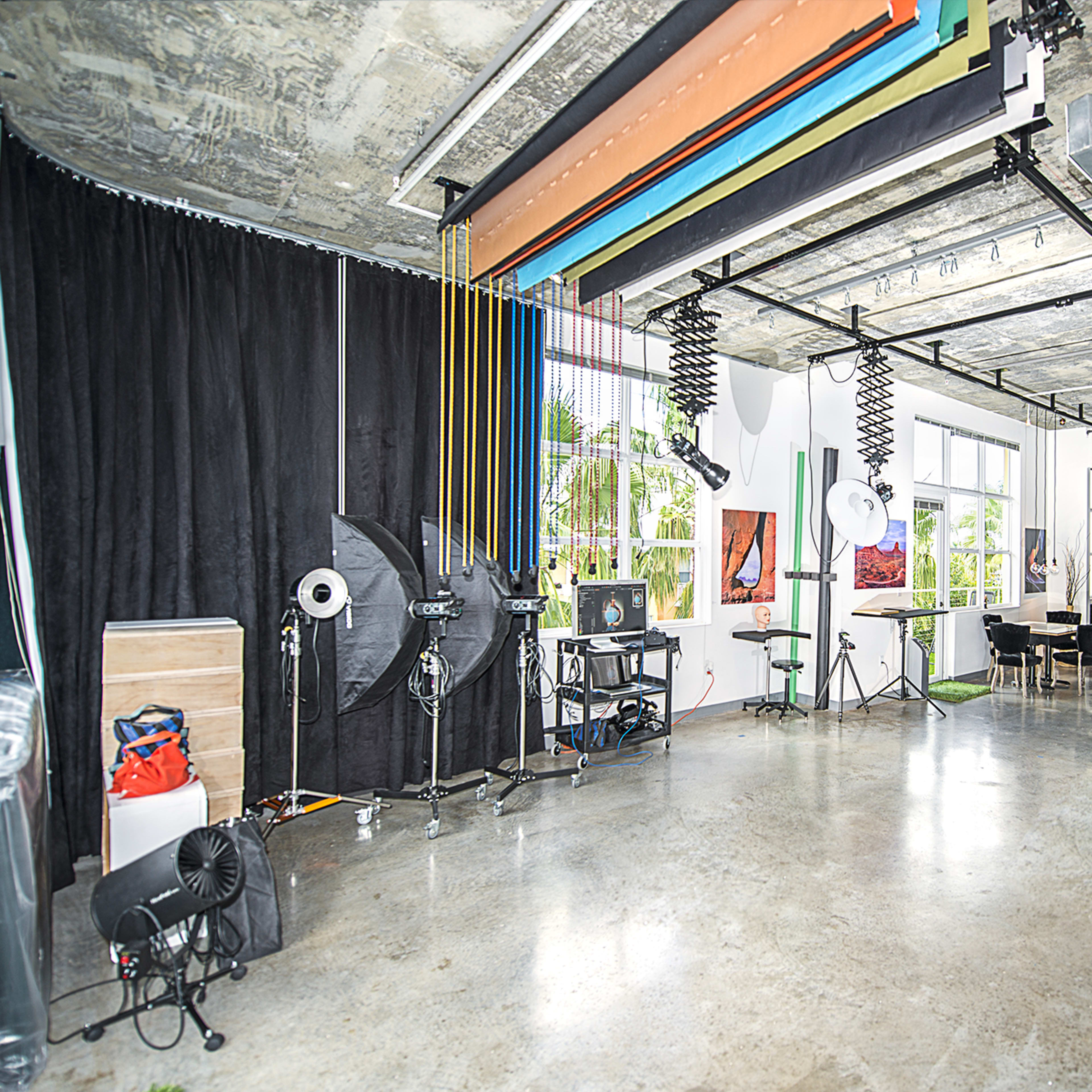 A spacious, modern studio with various lighting equipment, a seating area, and colorful backdrops hanging from the ceiling.