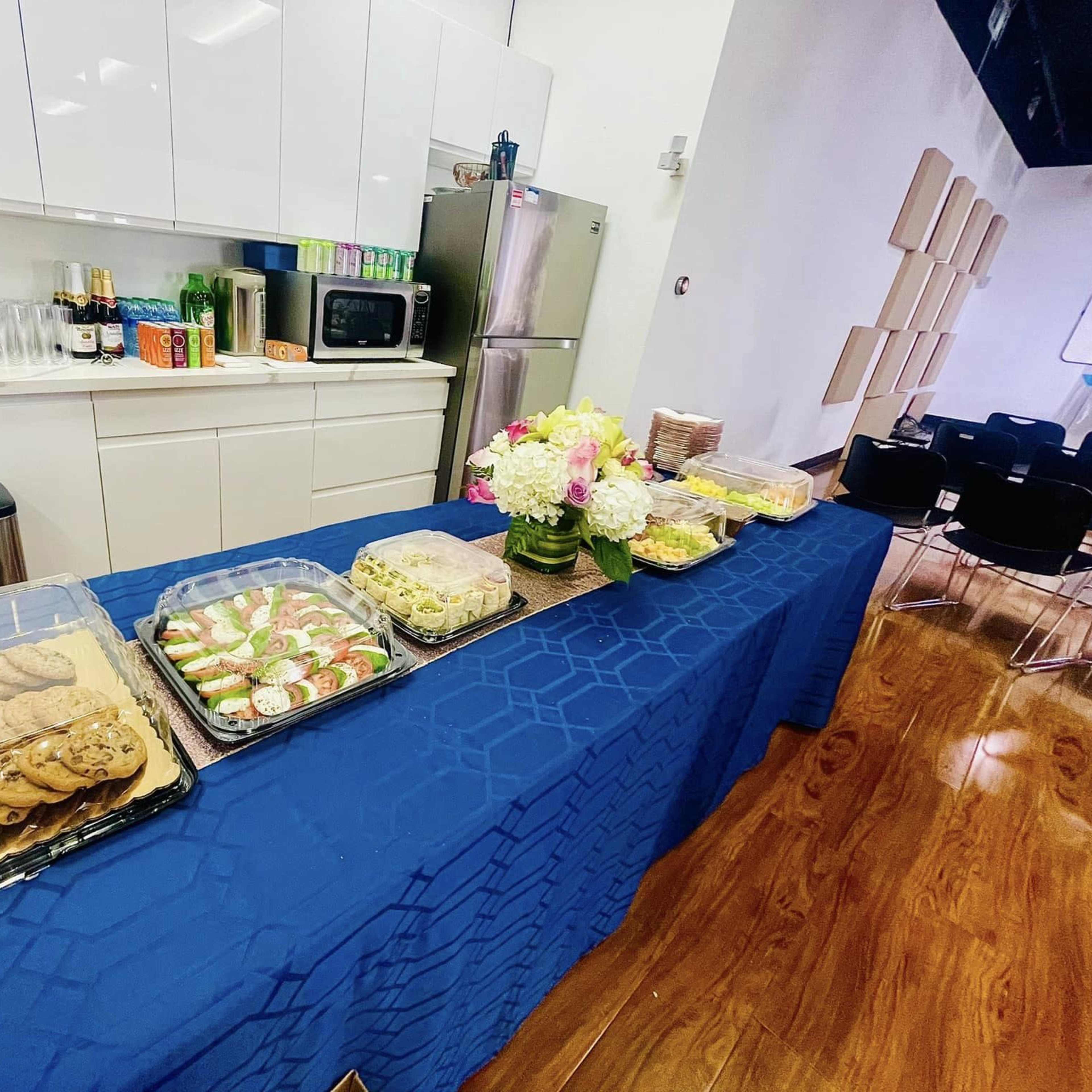A table covered with a blue cloth displays an assortment of food items, including cookies, sushi, and pasta dishes, in a modern kitchen setting.