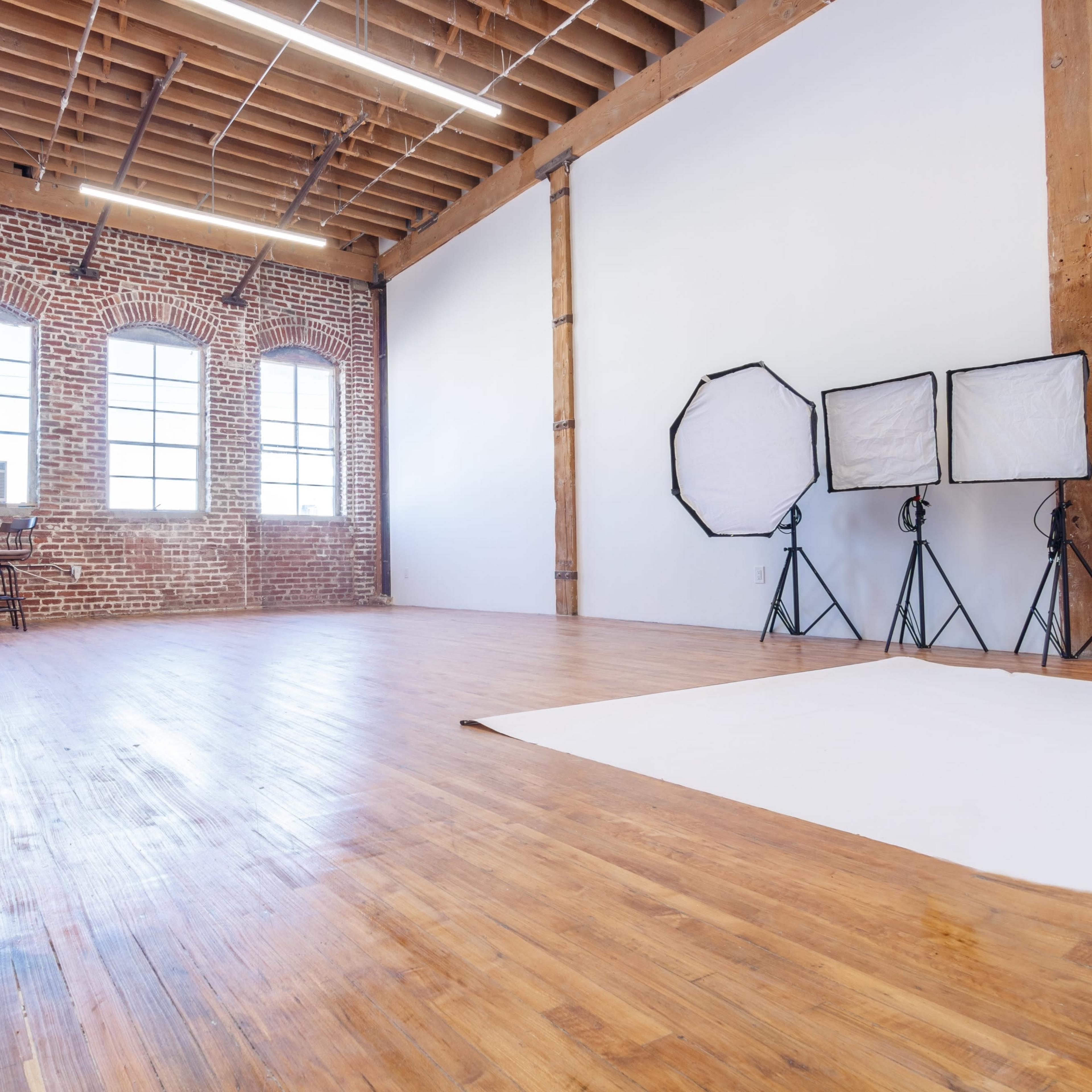 A spacious photography studio with wooden floors, exposed brick walls, and lighting equipment in the corner.