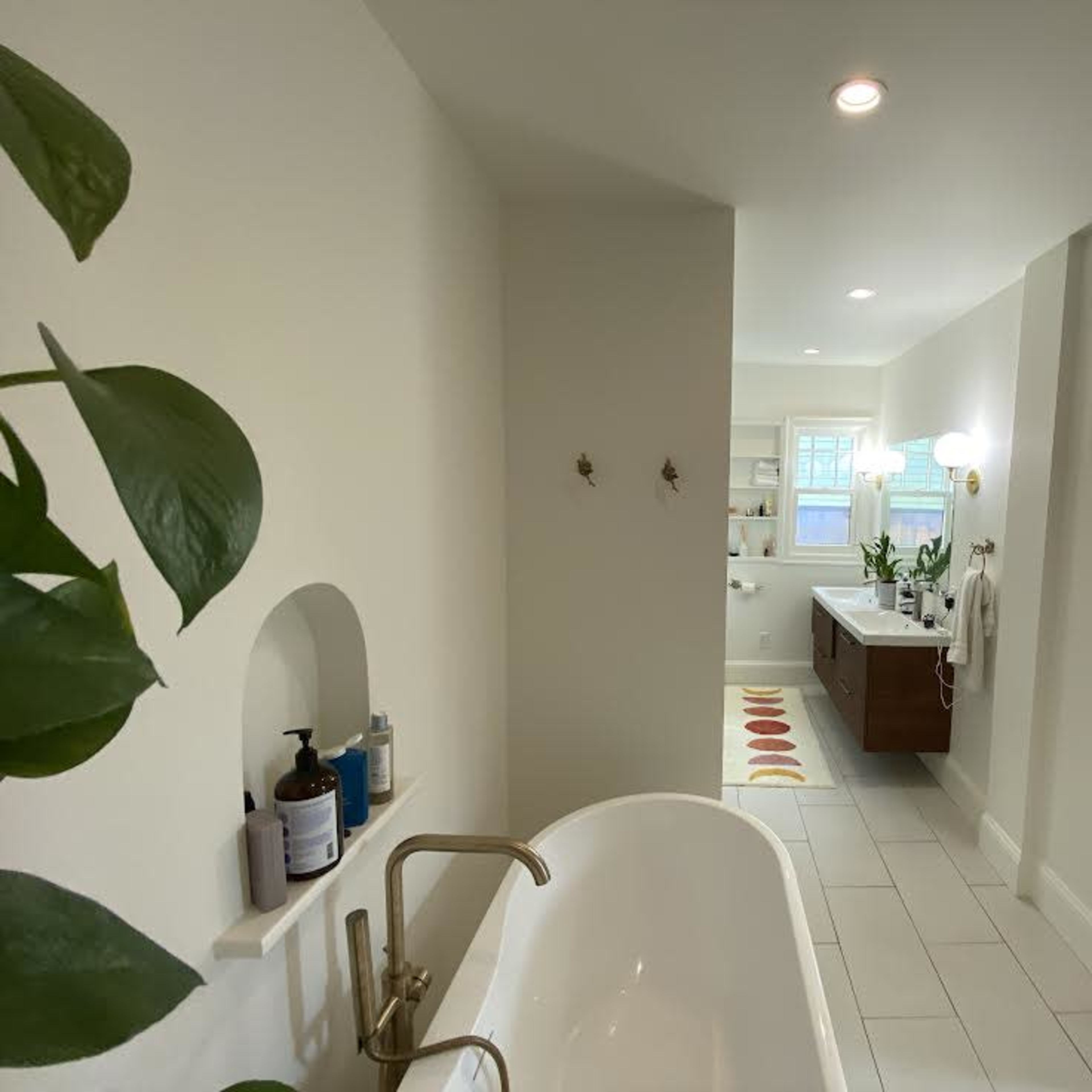 A bathroom with a freestanding tub, a wooden vanity, and a glimpse of a bedroom in the background.
