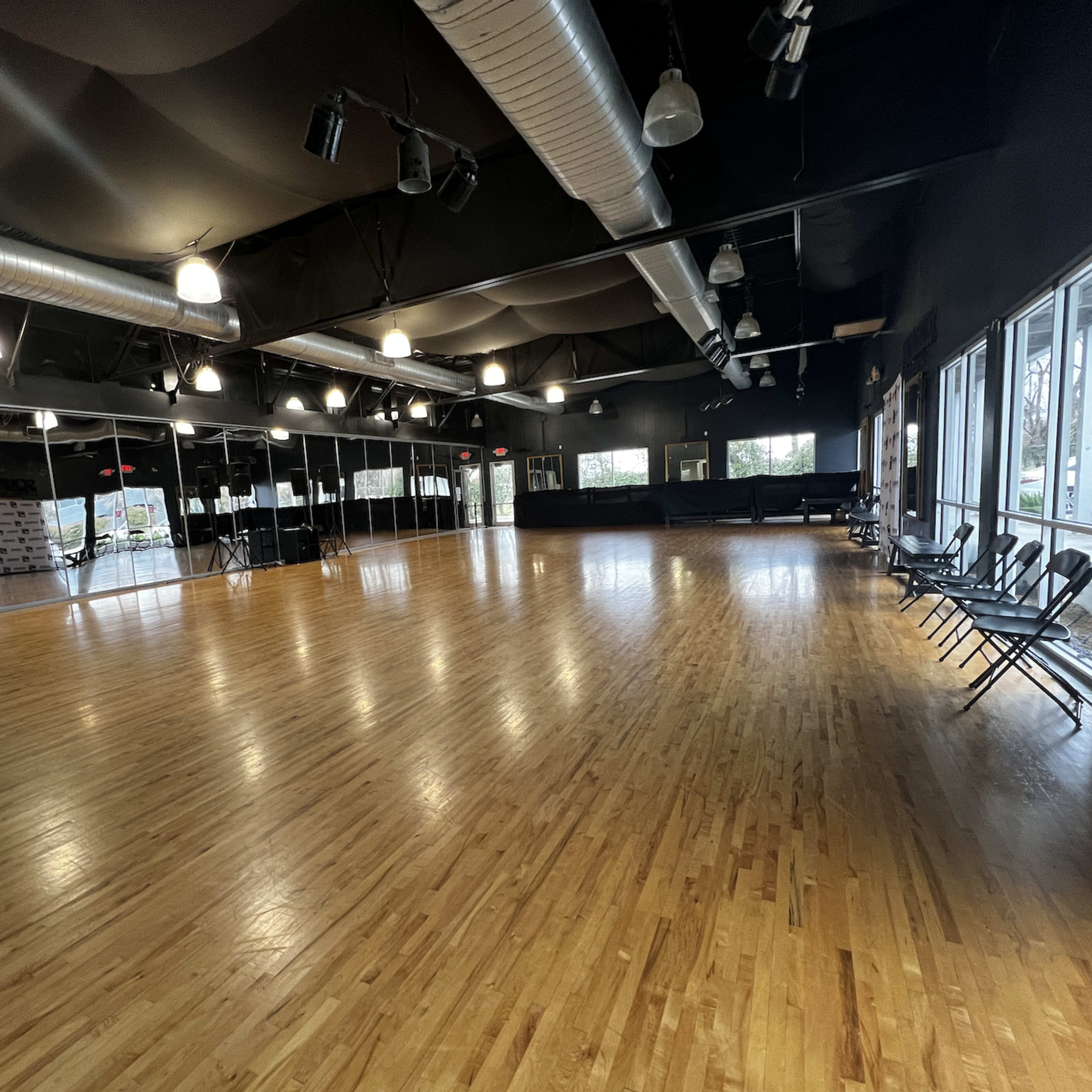 A spacious dance studio with polished wooden floors, black walls, and large windows allowing natural light to enter.