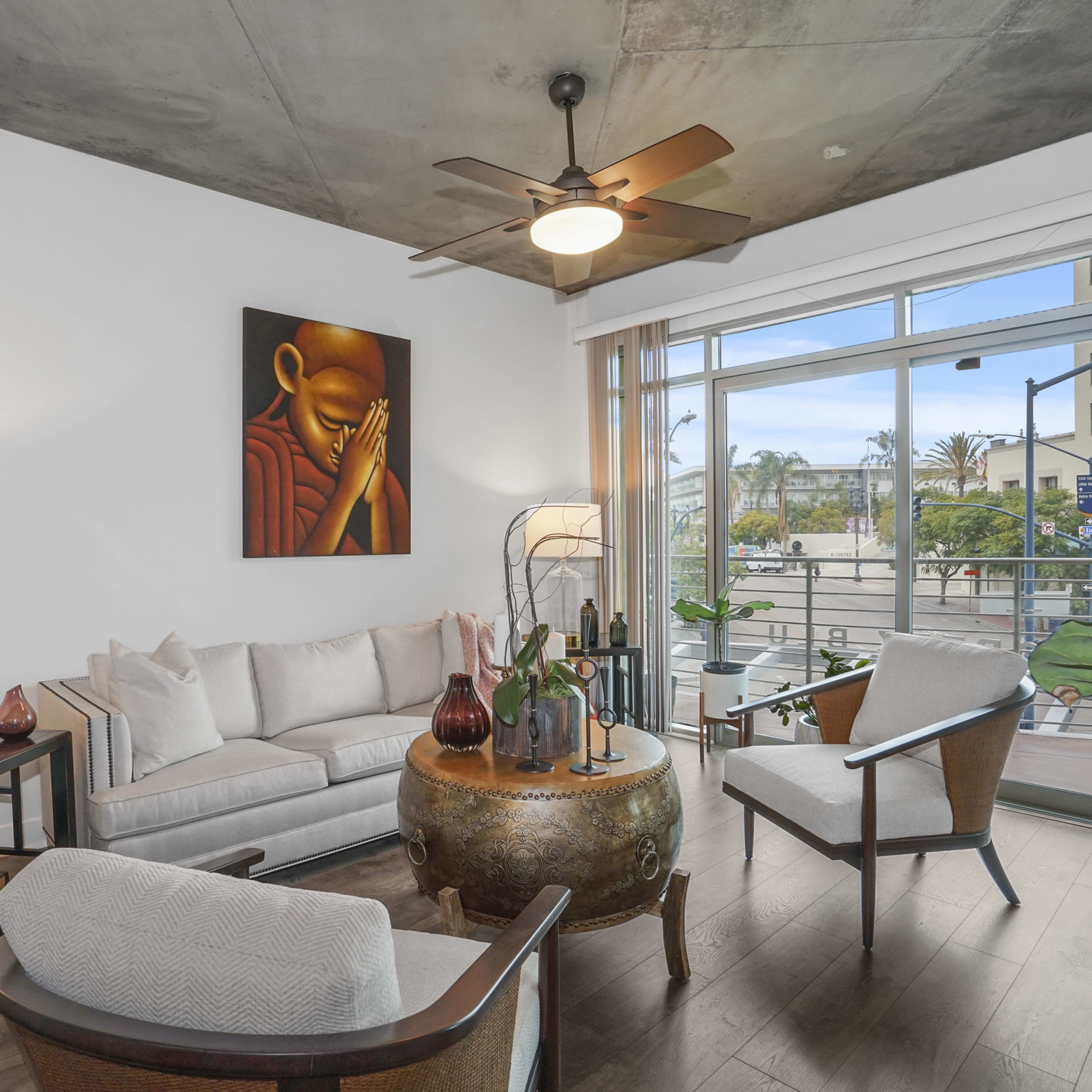 A modern living room features a light-colored sofa, wooden chairs, a round coffee table, and large windows with views of an outdoor setting.