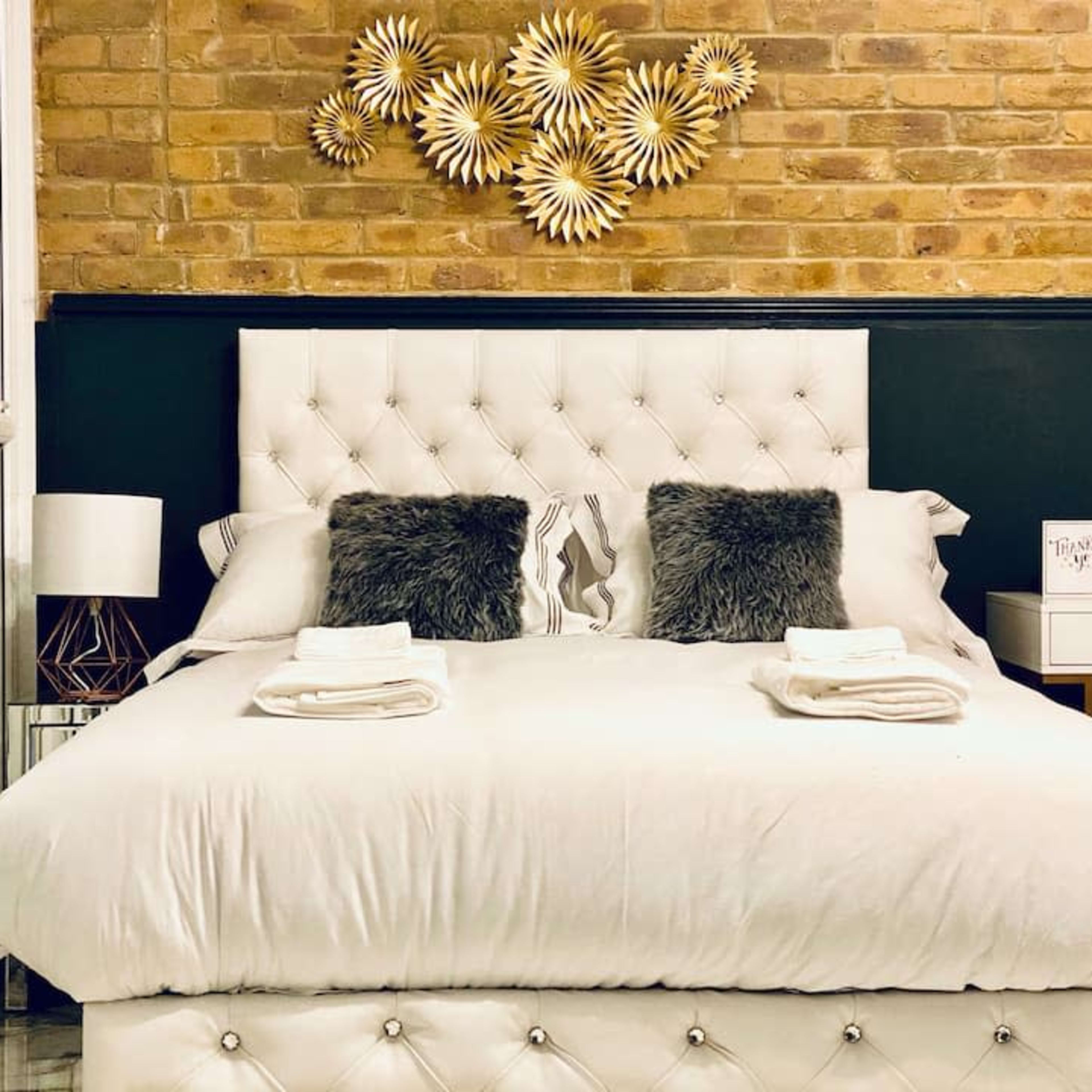 A neatly arranged bedroom features a tufted white bed with two decorative pillows, a bedside table, and a wall adorned with a sunburst art piece.