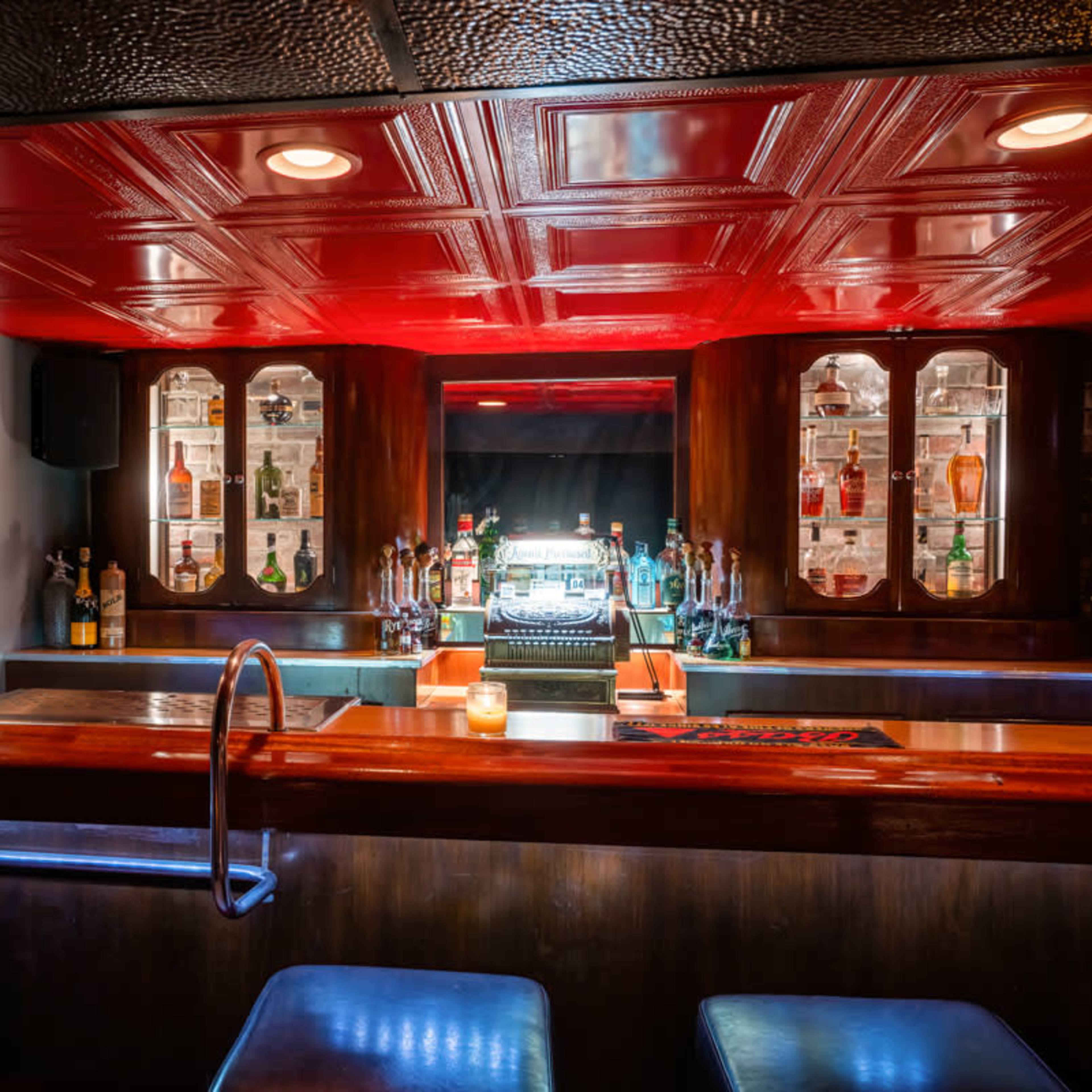 A well-lit bar with a wooden counter, illuminated bottles on display, and a red ceiling.