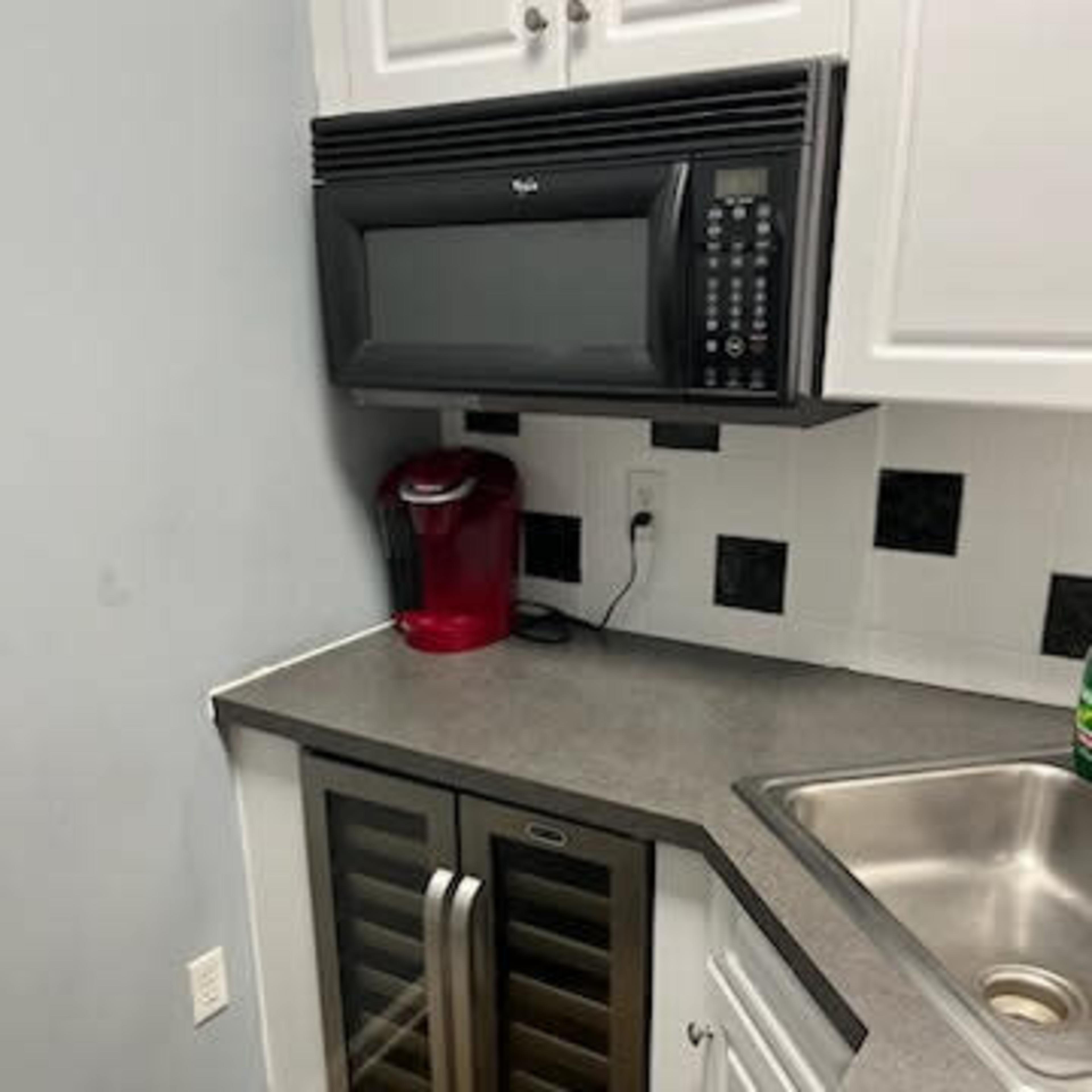 A corner of a kitchen with a black microwave mounted above a countertop that features a wine cooler beneath it and a sink nearby.