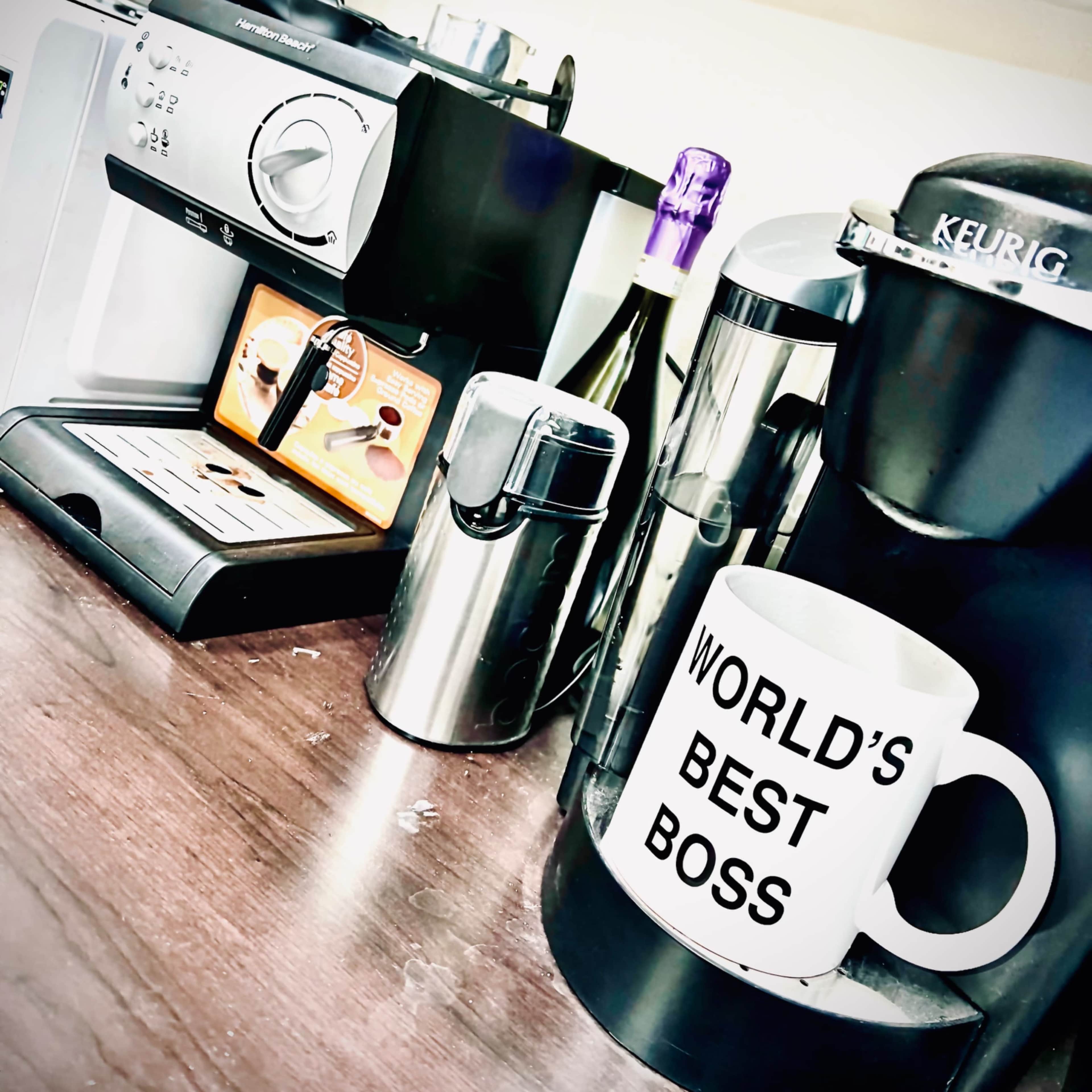 A coffee station features a Keurig machine, an espresso machine, a silver canister, and a large mug labeled "WORLD'S BEST BOSS."