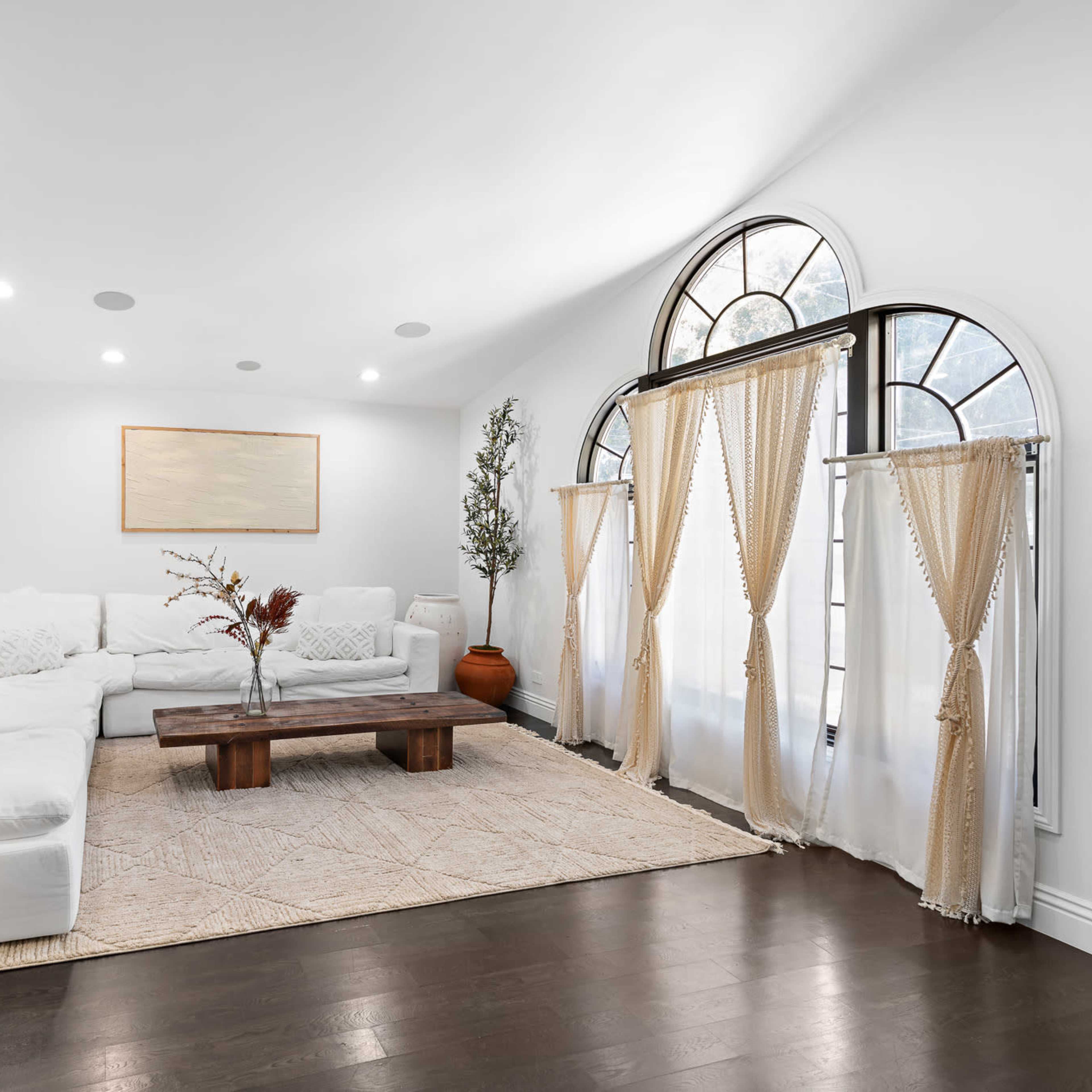 A bright living room features a large, white sectional sofa, a wooden coffee table, and arched windows draped with sheer curtains.
