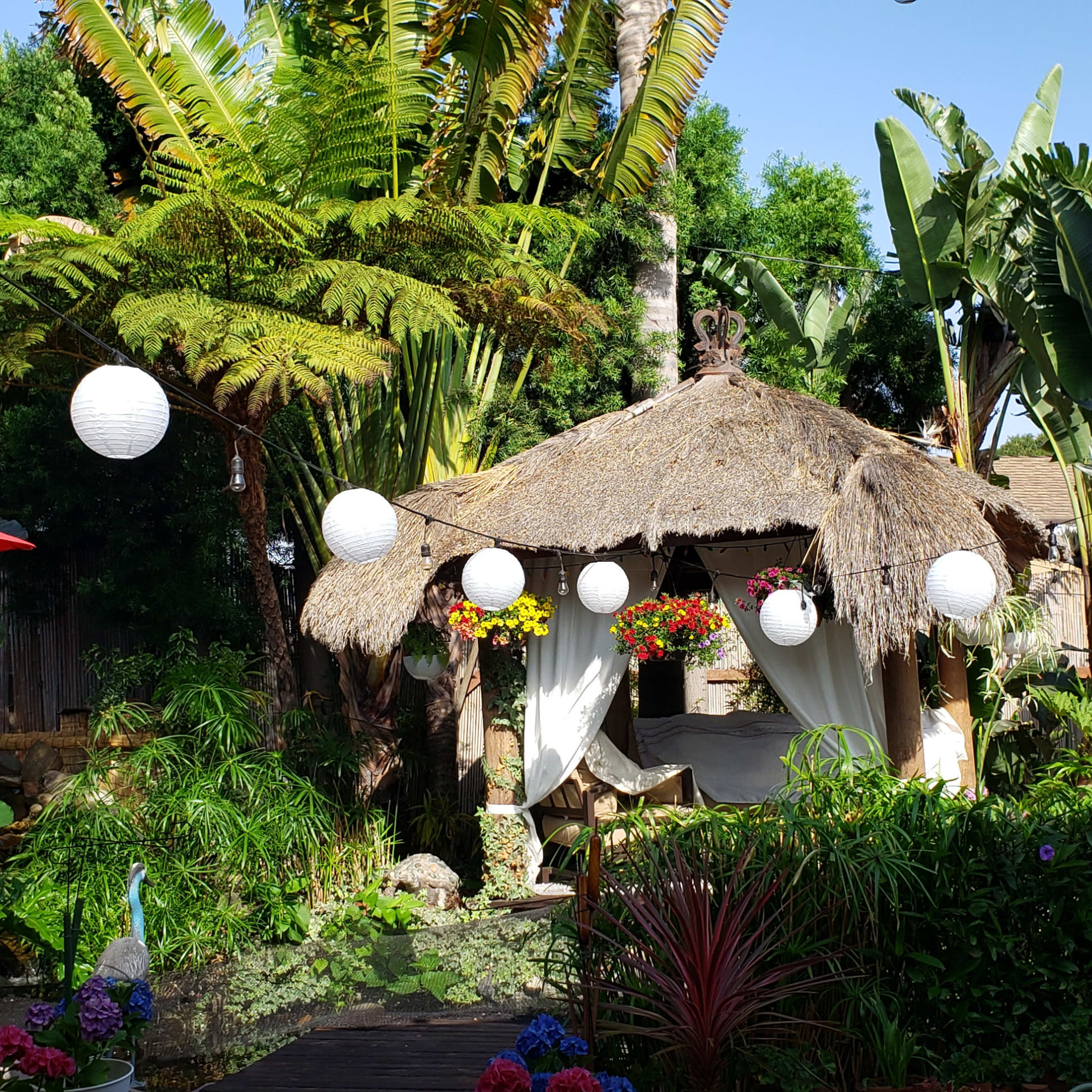 A vibrant garden with a thatched-roof gazebo surrounded by tropical plants and hanging lanterns.