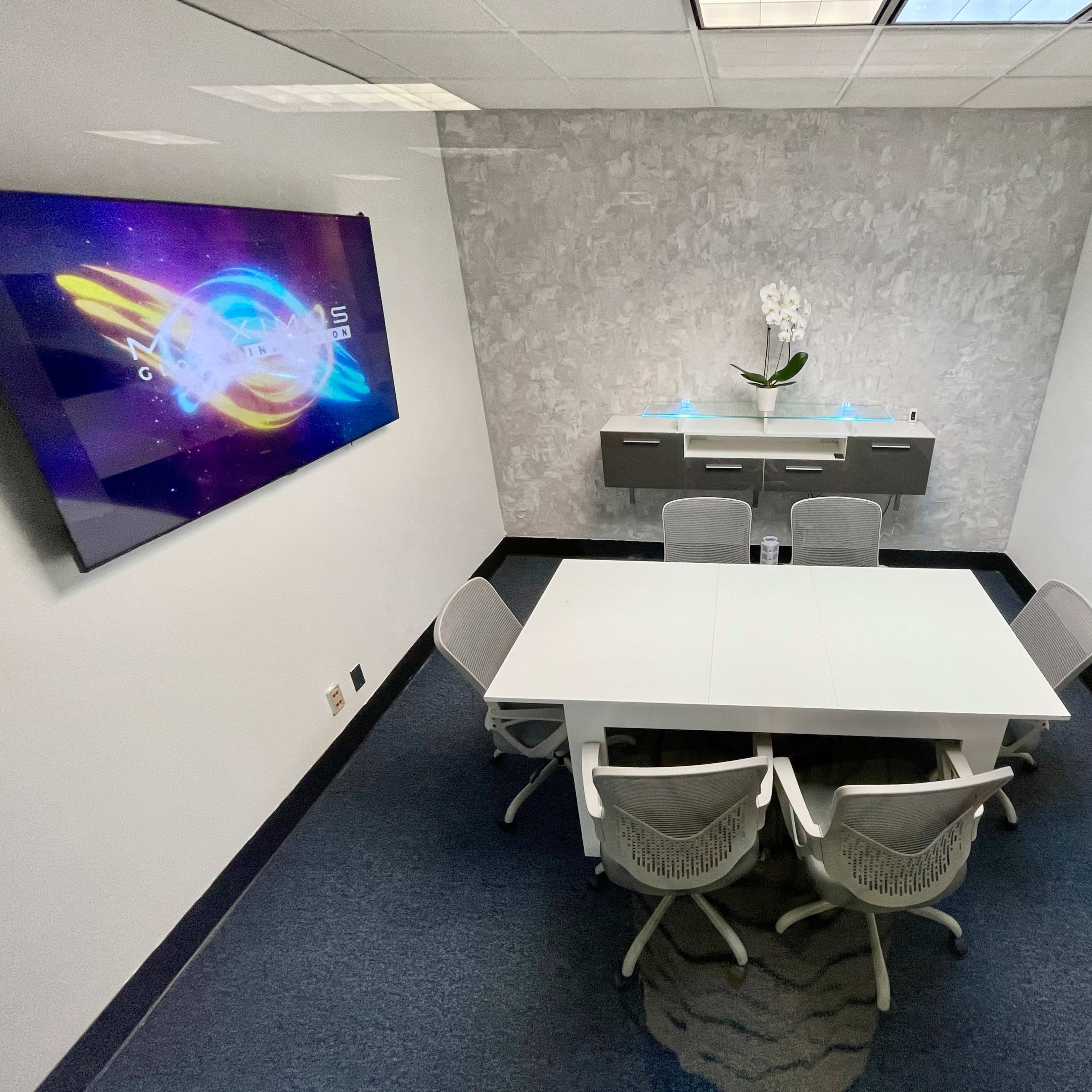 A conference room features a white table surrounded by six chairs, a mounted television, and a minimalist wall décor with an orchid centerpiece.