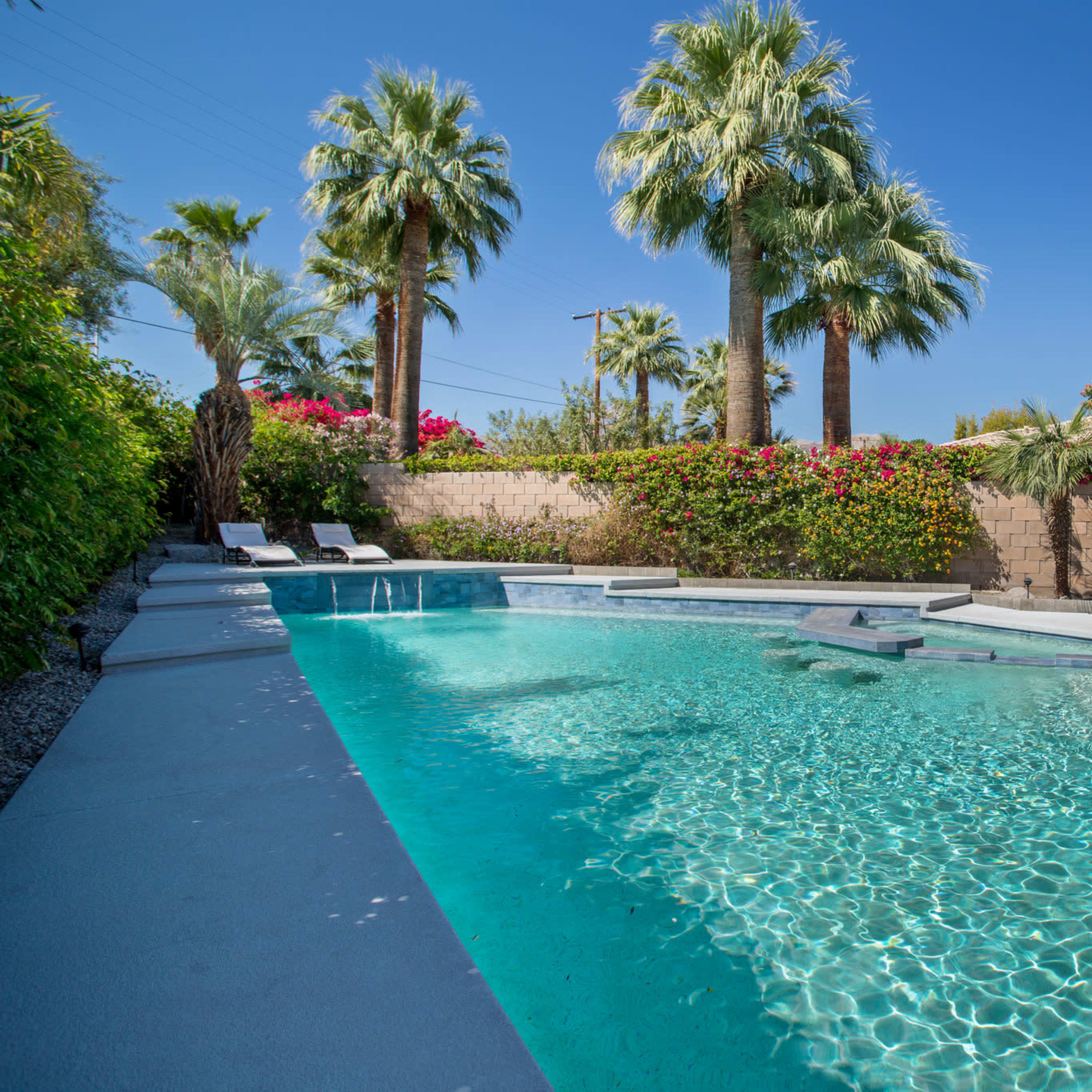 A clear swimming pool surrounded by palm trees and lush greenery, with several lounge chairs and a shaded seating area nearby.