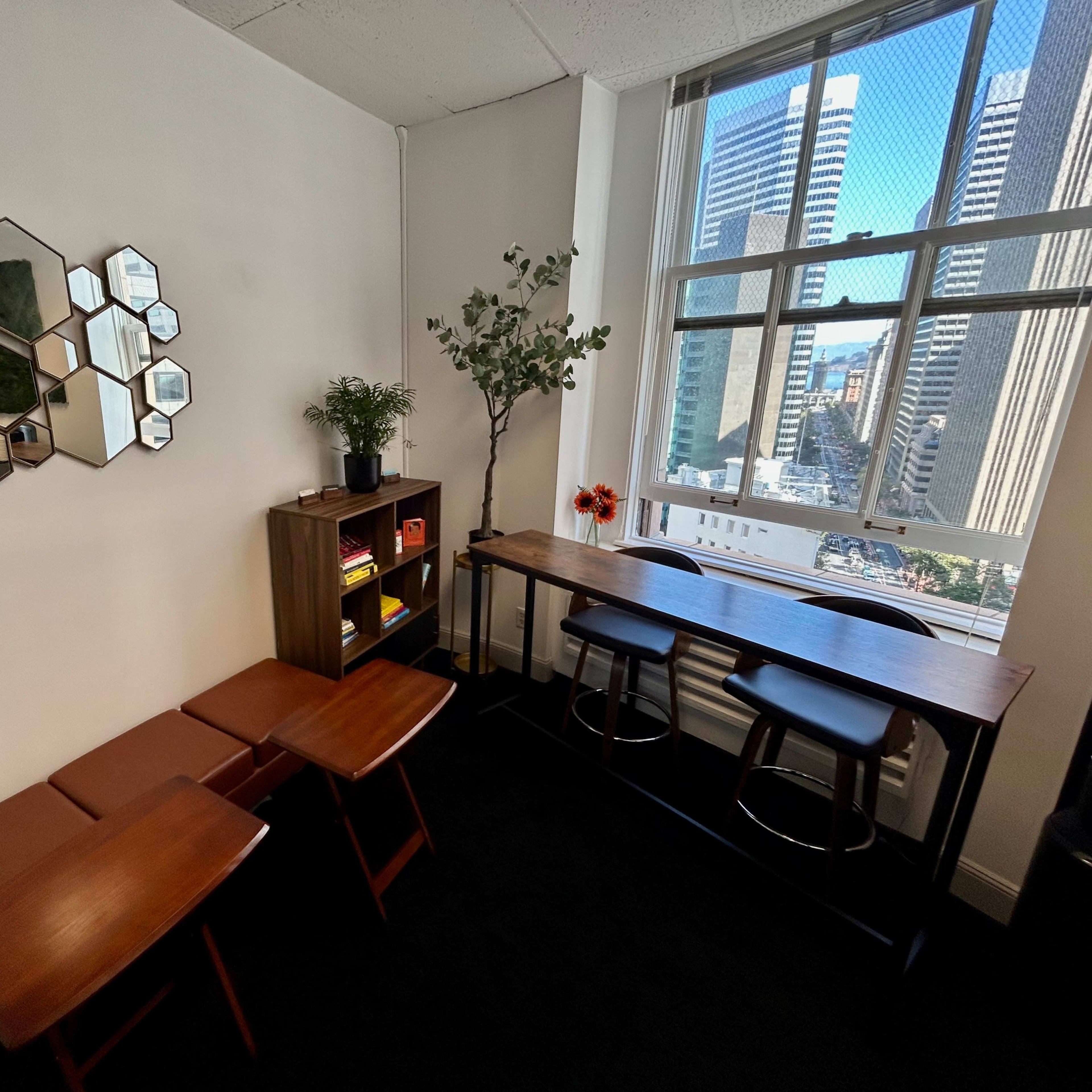 A small office space with a couple of wooden tables, a bookshelf, a vase with flowers, and a large window overlooking a city skyline.