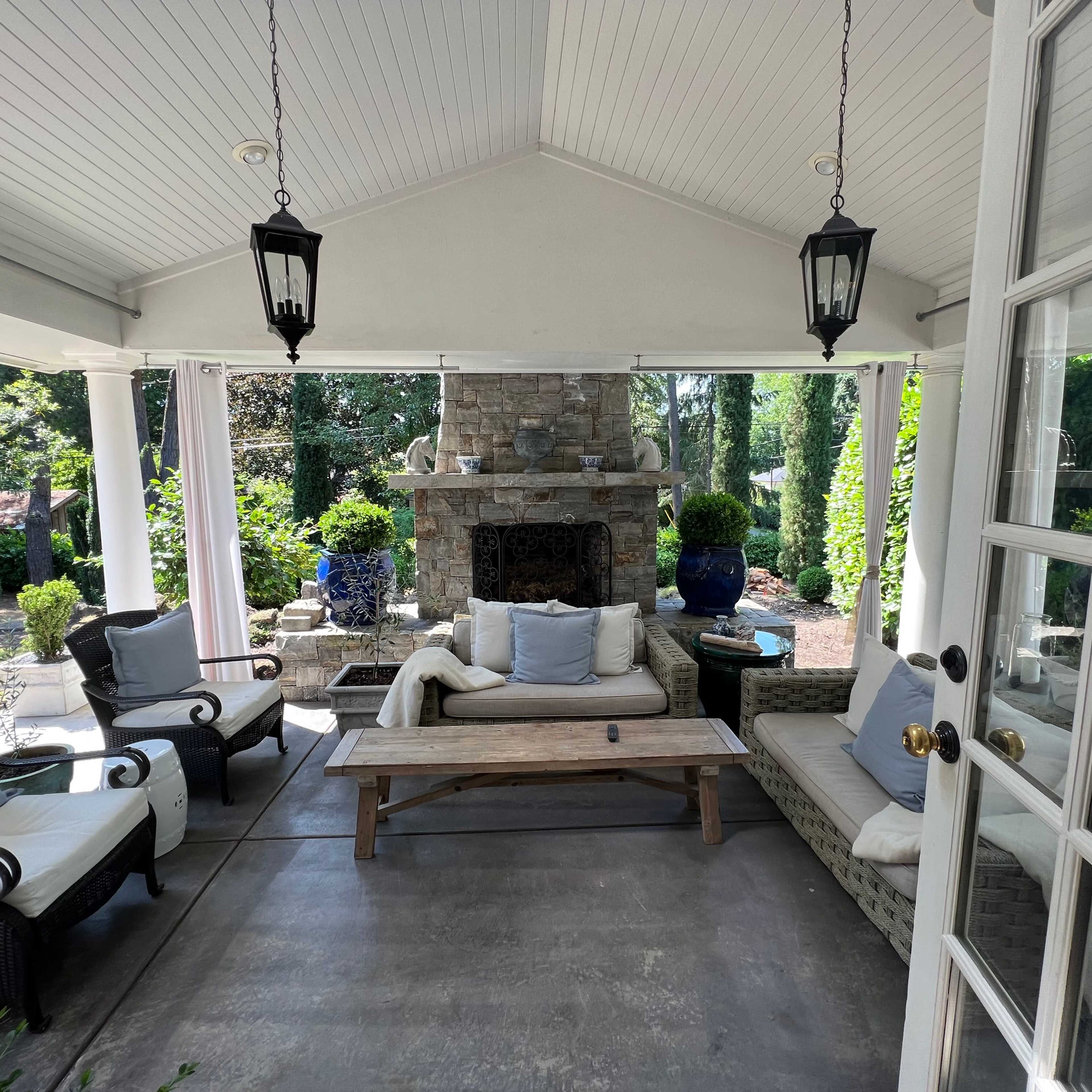 A covered outdoor sitting area with a stone fireplace, wicker furniture, and lush greenery in the background.