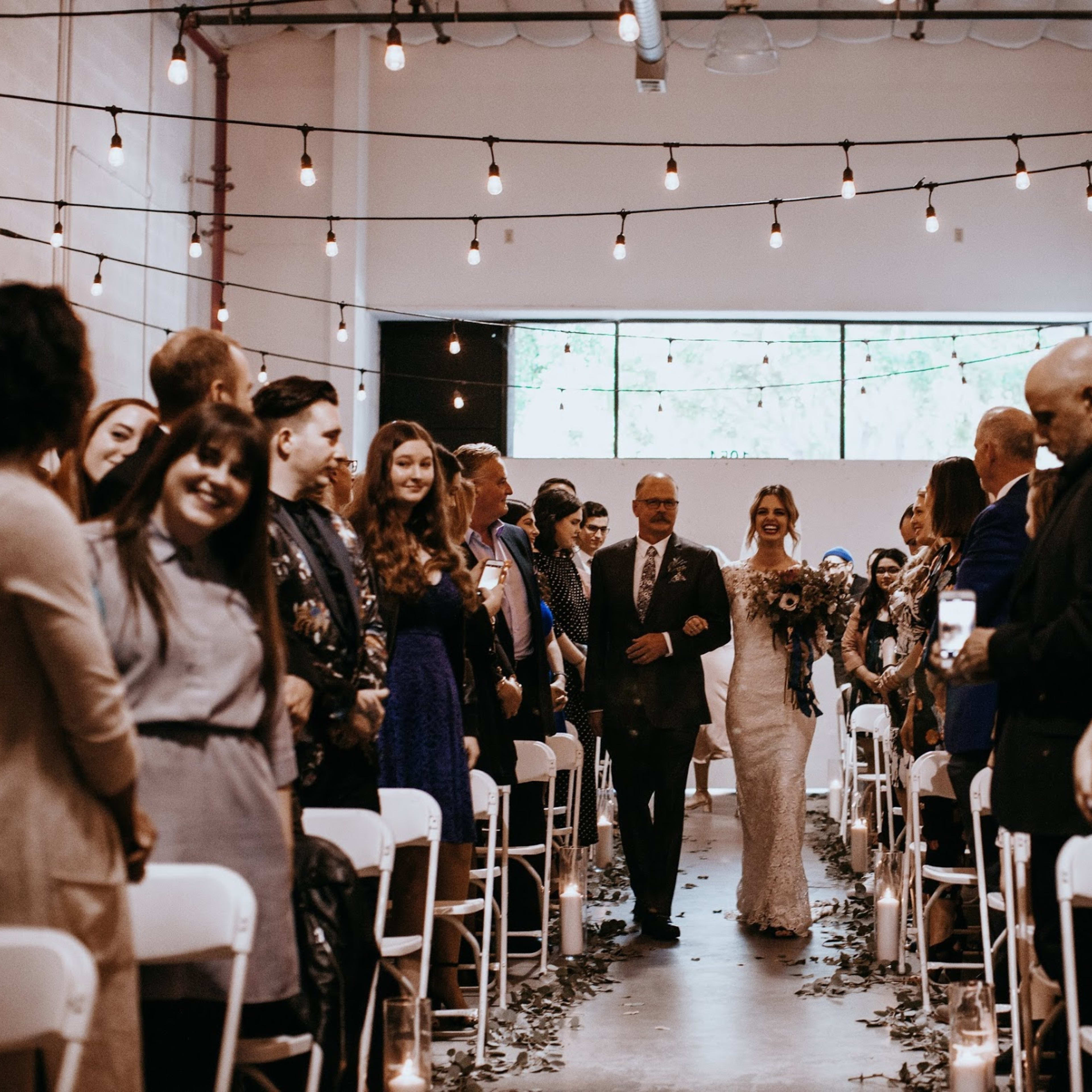 A bride walks down the aisle accompanied by her father, with guests seated on either side under string lights in a decorated venue.