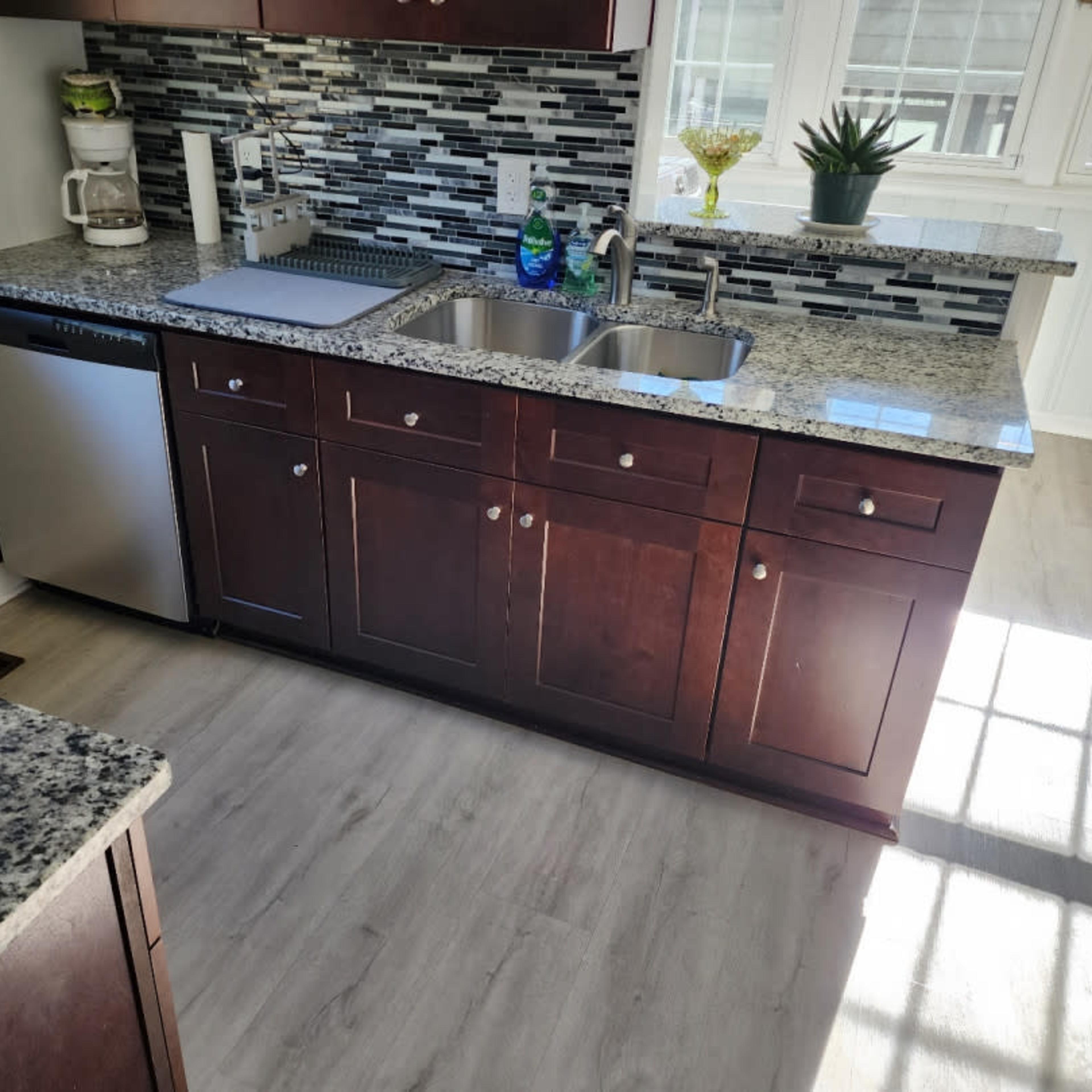 A kitchen with dark wood cabinets, a stainless steel dishwasher, a double sink, and a countertop made of speckled granite, illuminated by natural light.
