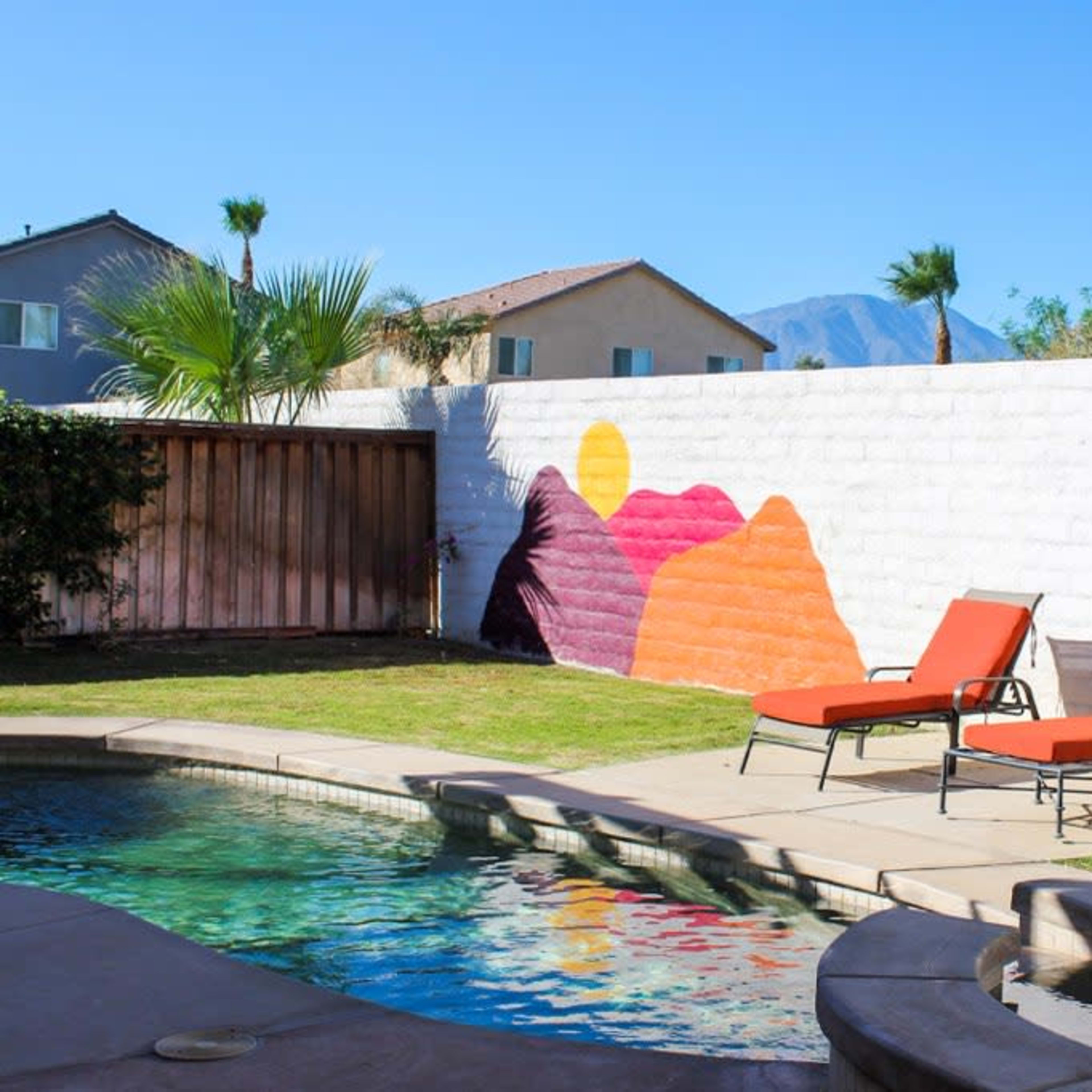A swimming pool area with two orange lounge chairs beside a colorful mountain mural on a white wall.