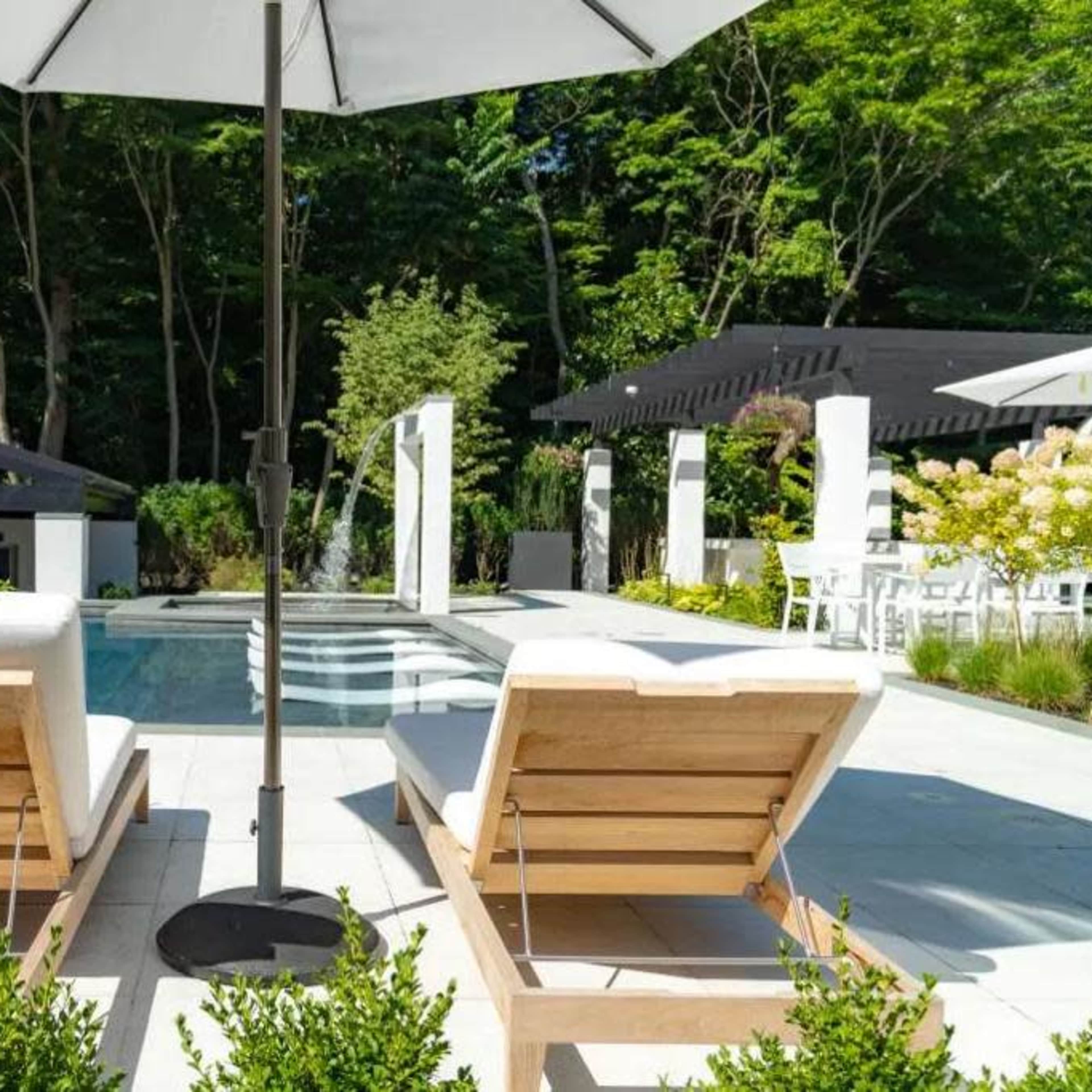 Two lounge chairs under a large umbrella beside a swimming pool, surrounded by greenery and garden landscaping.