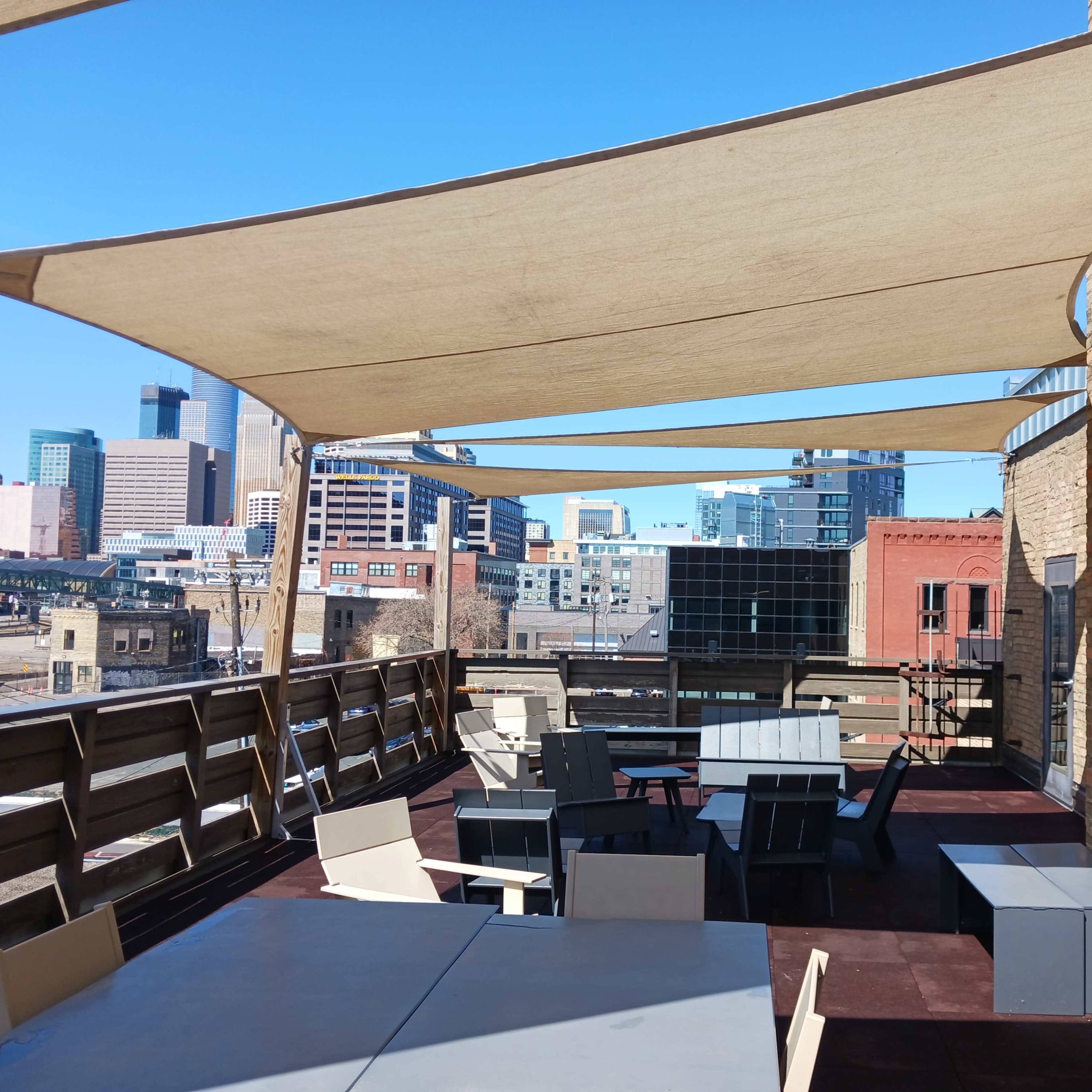 A rooftop terrace features several tables and chairs shaded by large fabric canopies, with a city skyline visible in the background.