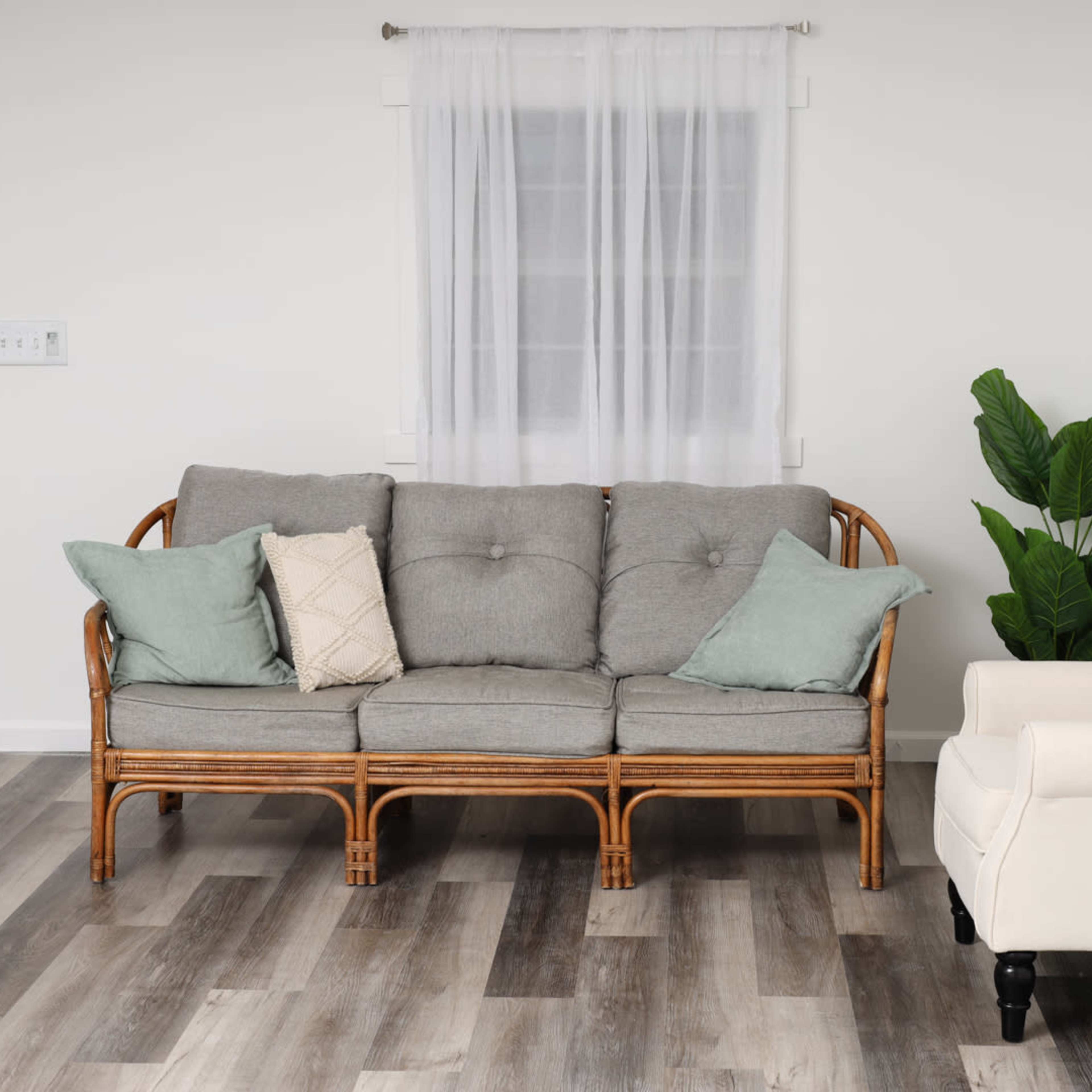 A gray sofa with cushions sits against a white wall, beside a light-colored armchair and a potted plant, with a window and door visible in the background.