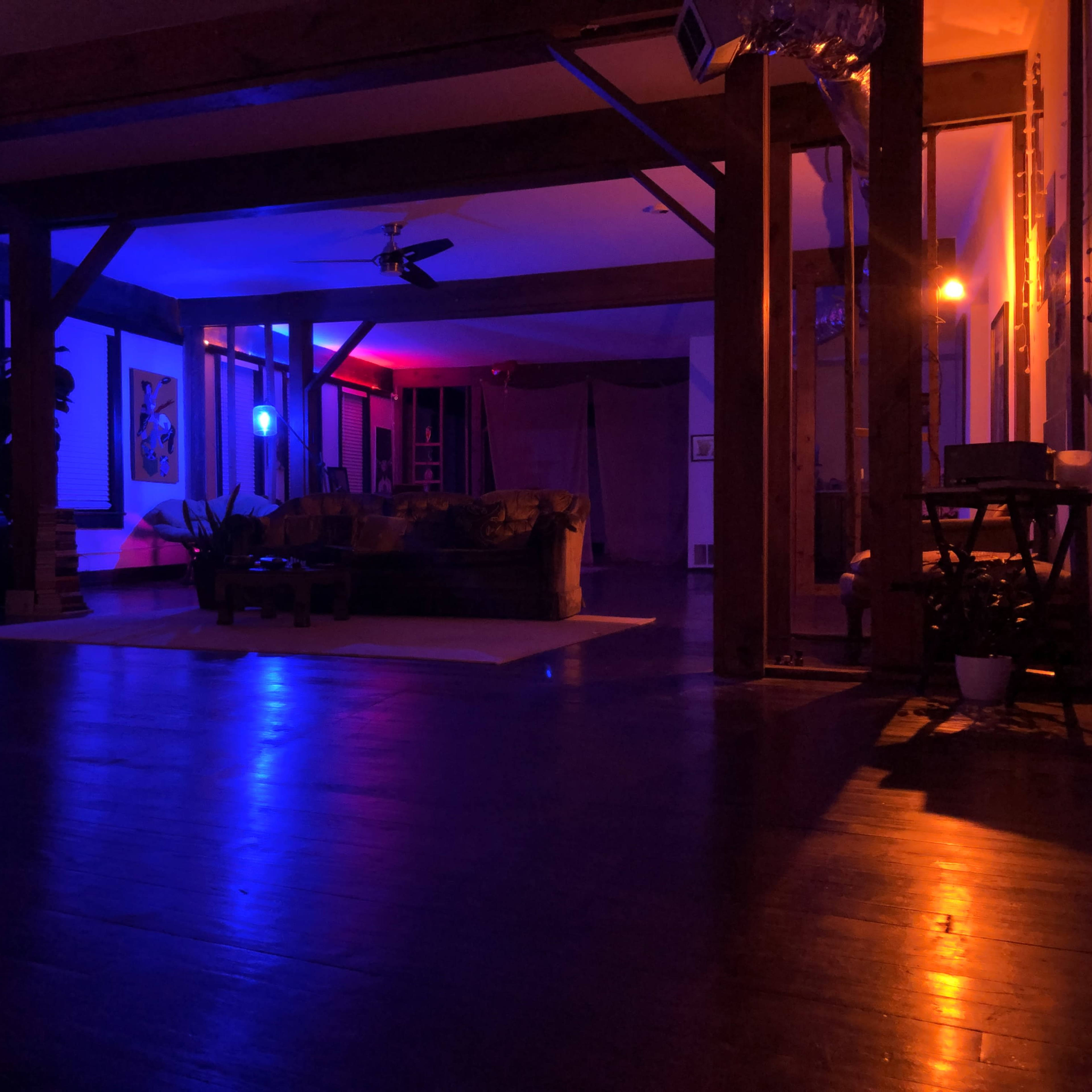 A dimly lit interior space with colorful lighting, featuring a couch, artwork on the walls, and various furnishings.