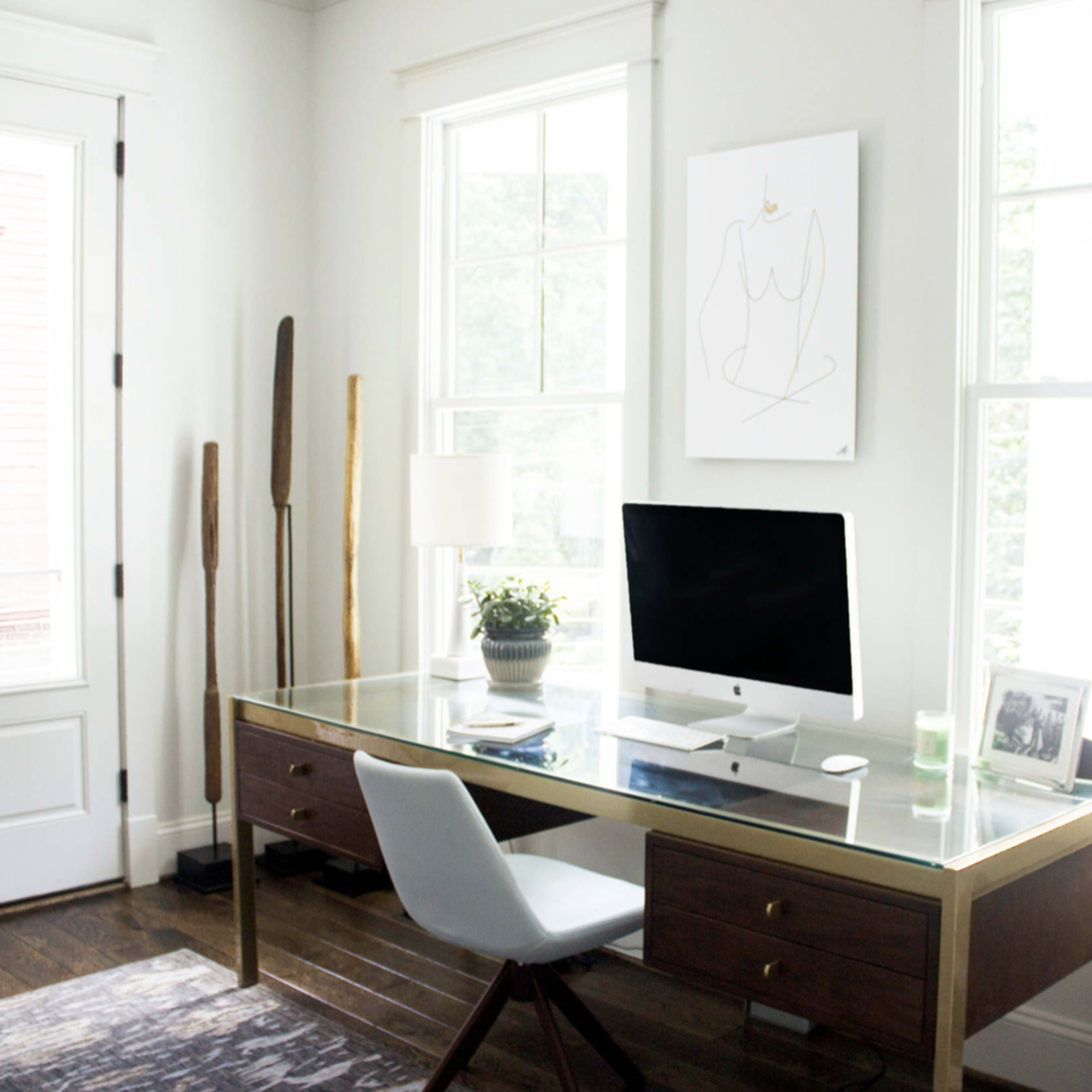 A modern home office features a glass-top desk with an iMac, a potted plant, and natural light streaming through large windows.