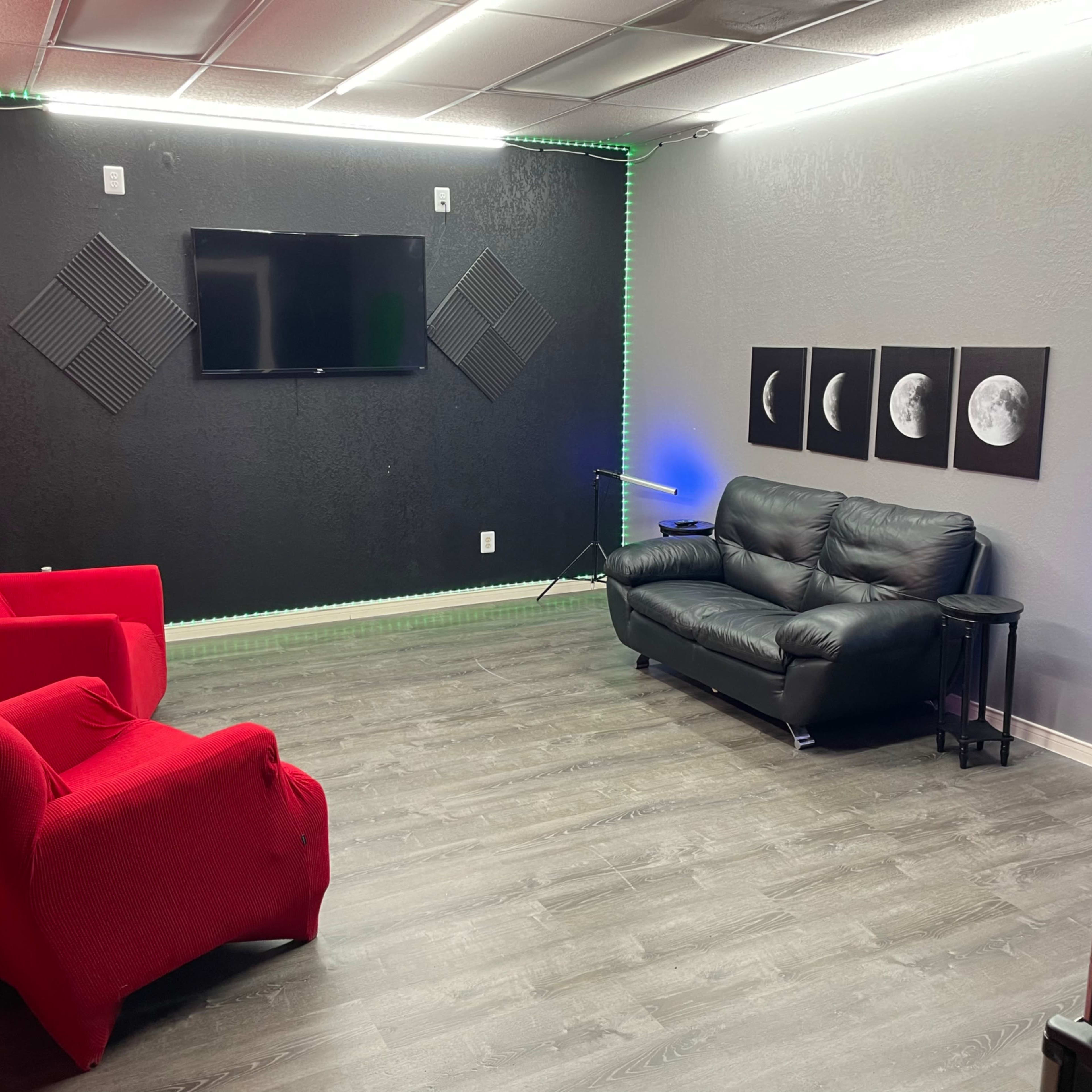 The room features a television mounted on the wall, a black sofa, two red chairs, and three framed lunar images.