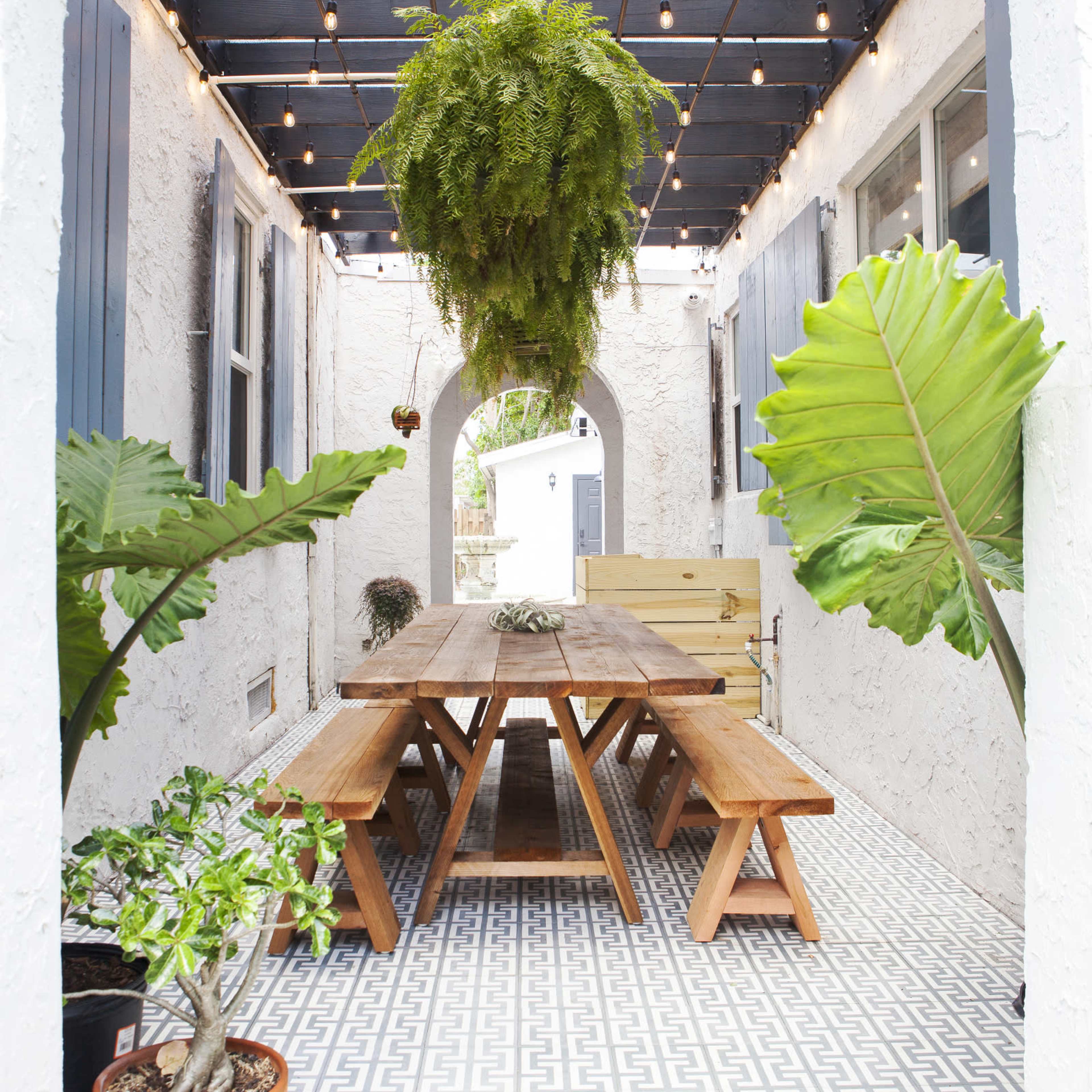 A patio features a wooden dining table surrounded by benches under a trellised roof with hanging plants and string lights.