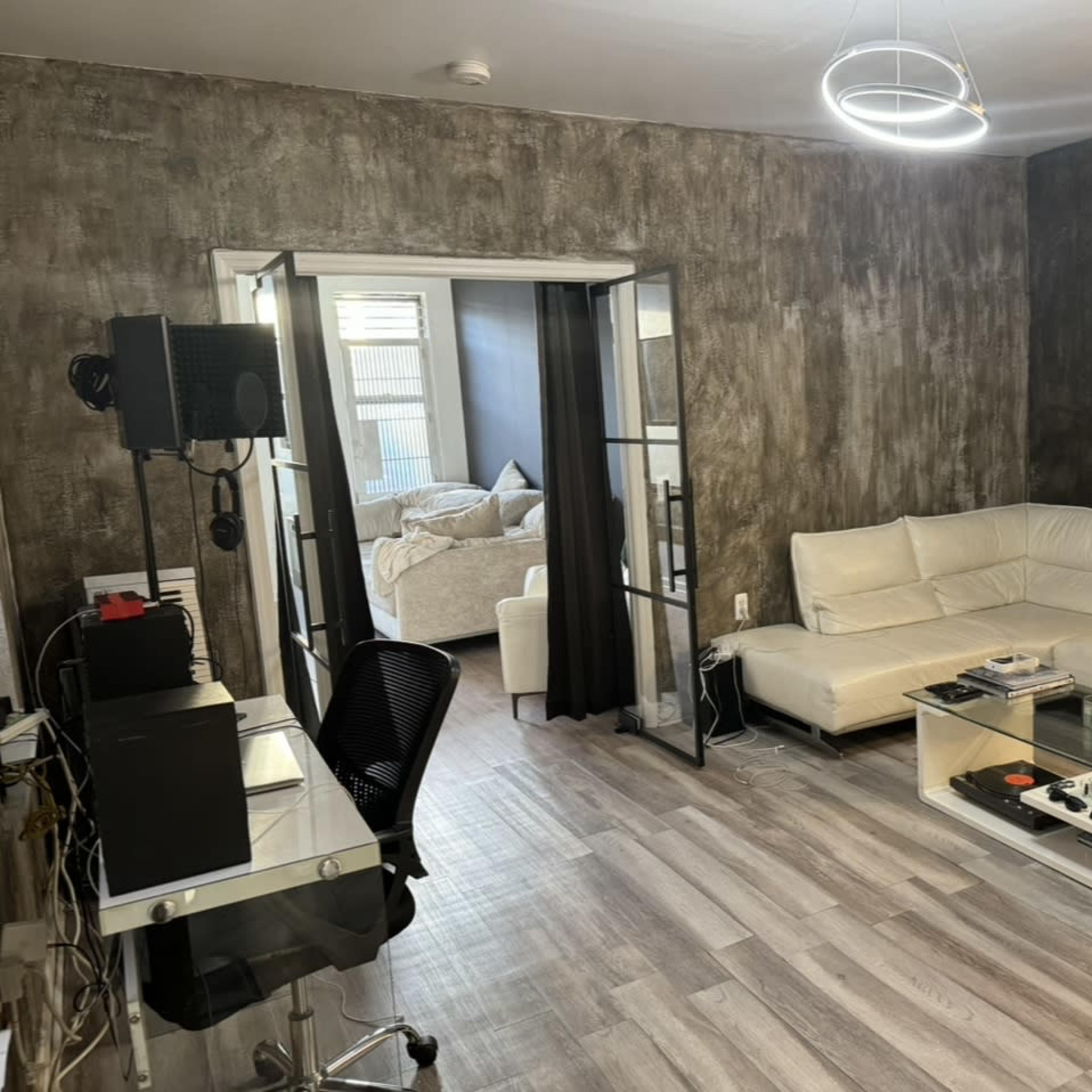 A modern living room with a white sectional sofa, a glass coffee table, and a desk with a computer setup, leading to an adjoining bedroom through glass doors.