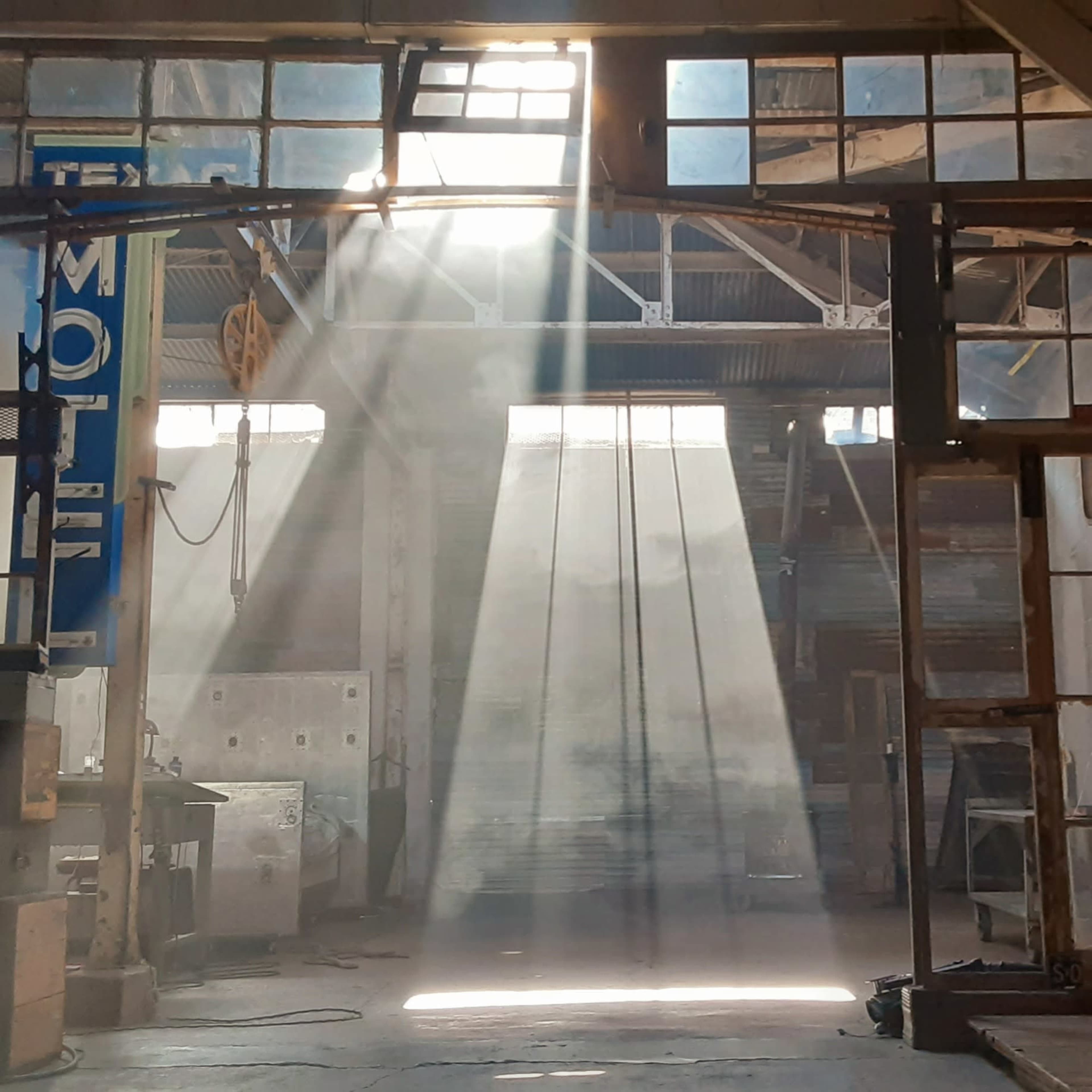 Sunlight streams through the windows of an empty industrial warehouse, creating beams of light in the dusty air.