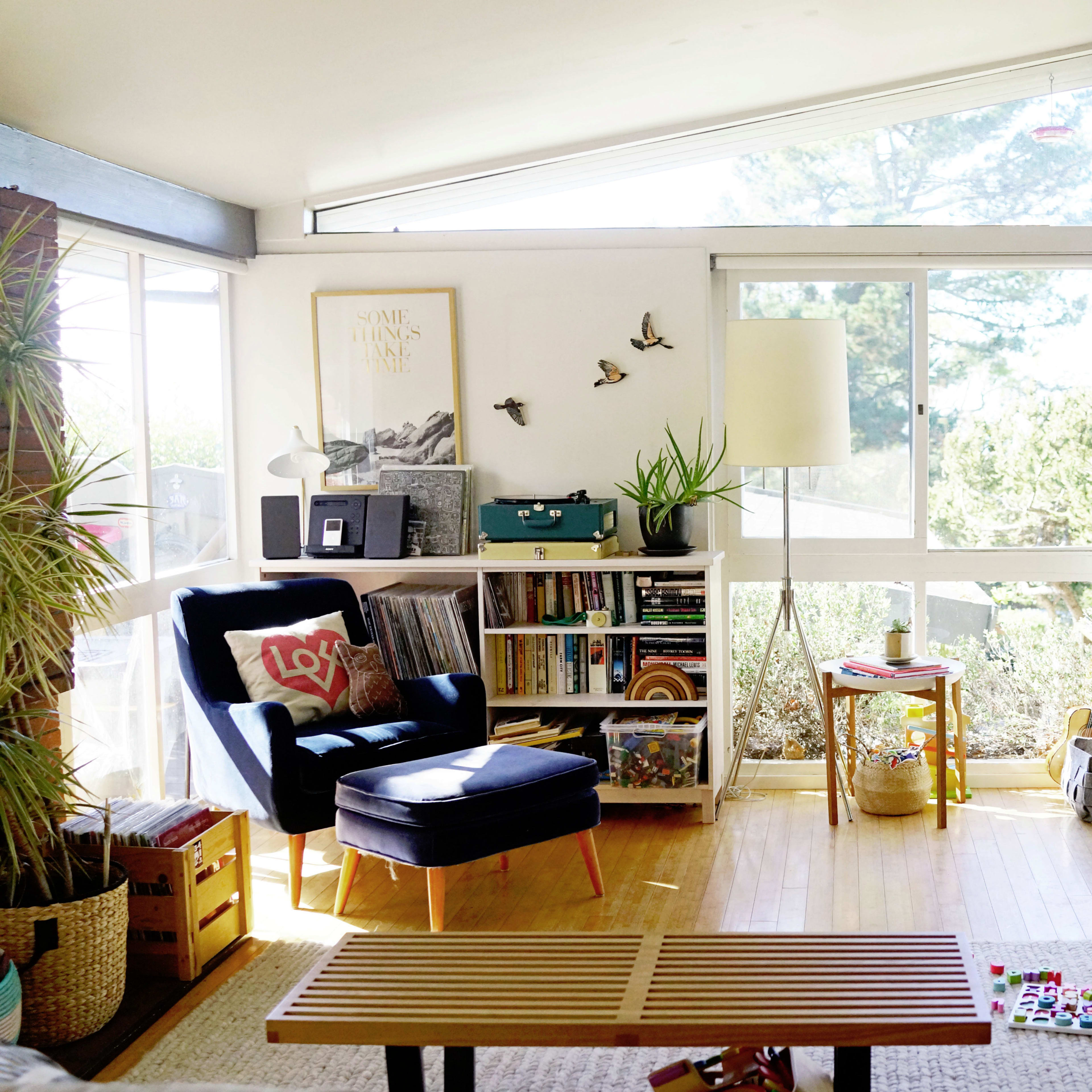 The living room features a large window with natural light, a blue armchair, a wooden coffee table, and various plants and decor items.