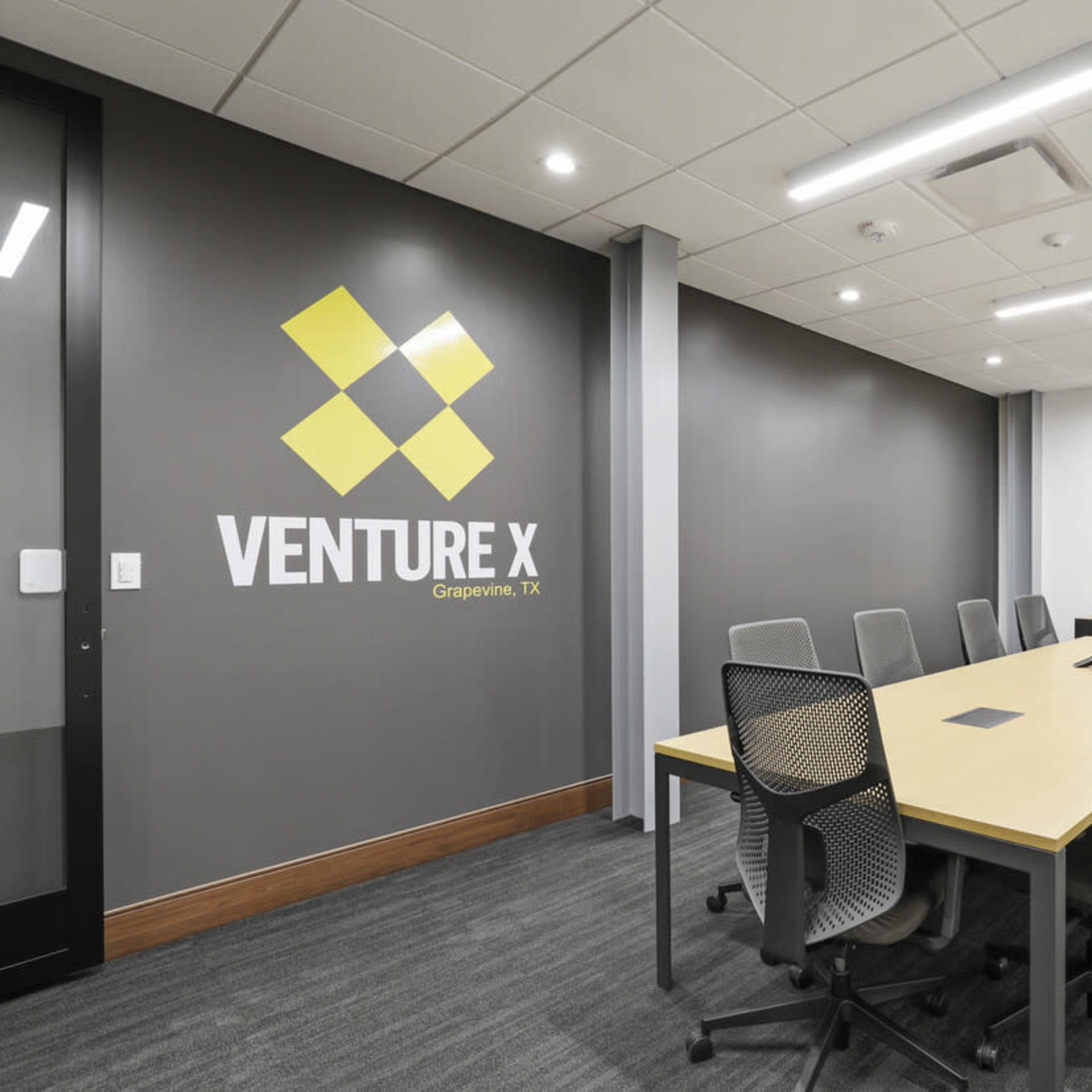 A modern meeting room with a long table, ergonomic chairs, a large screen, and a wall featuring the "VENTURE X" logo.