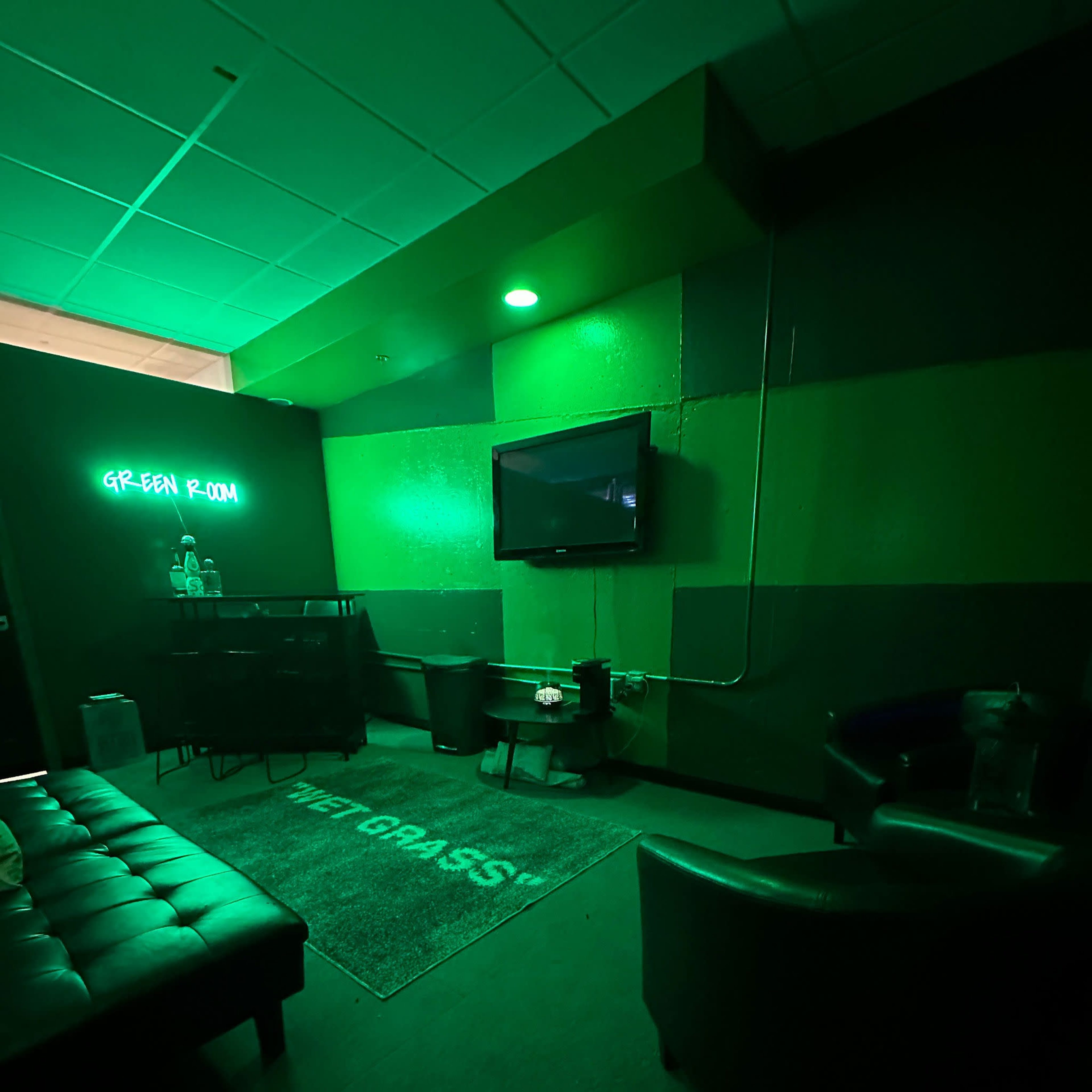 A dimly lit green room features a couch, two chairs, a television on the wall, and a neon sign.