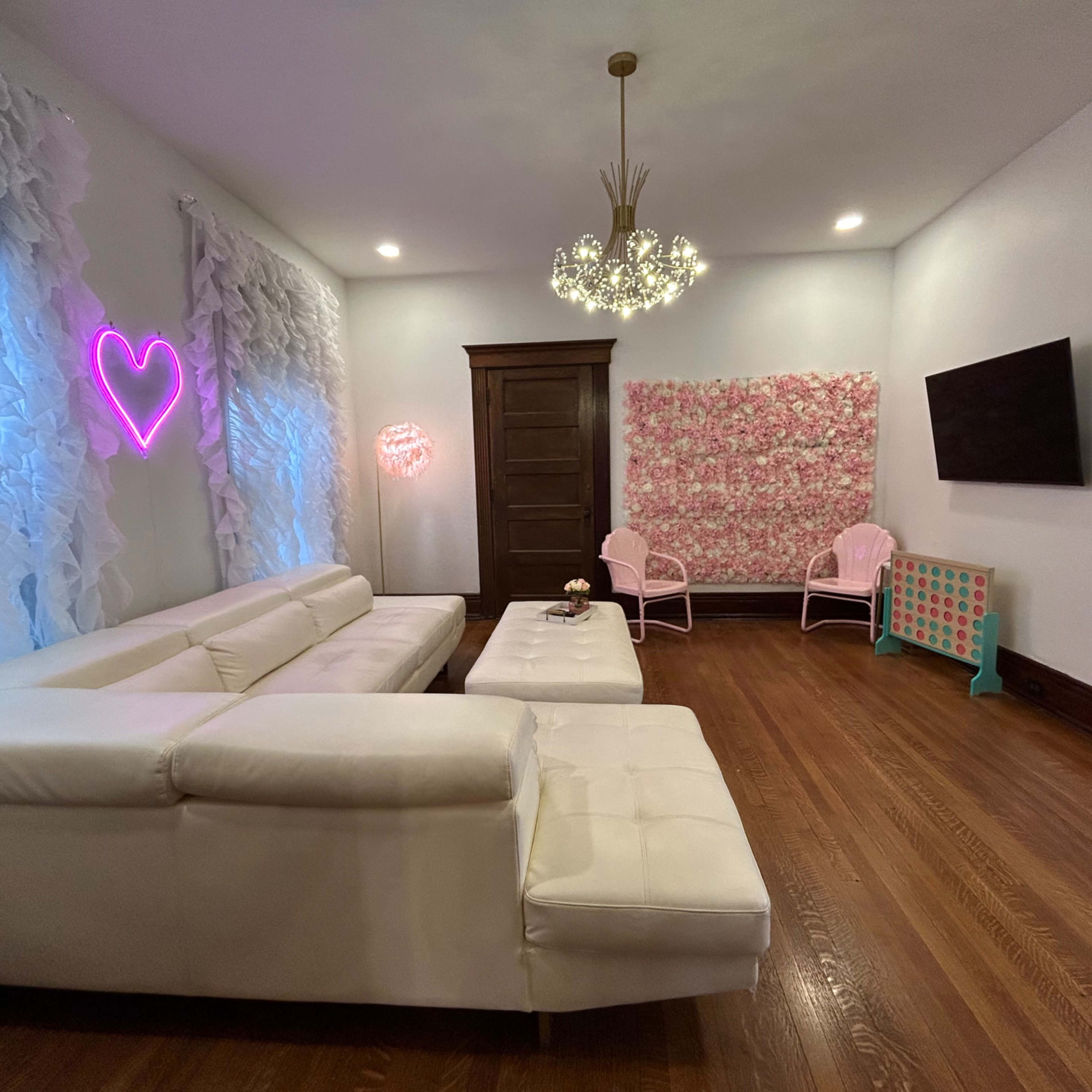 A modern living room with a white sectional sofa, a pink neon heart light, an ornate light fixture, and floral decor on one wall, along with a television mounted opposite the seating area.