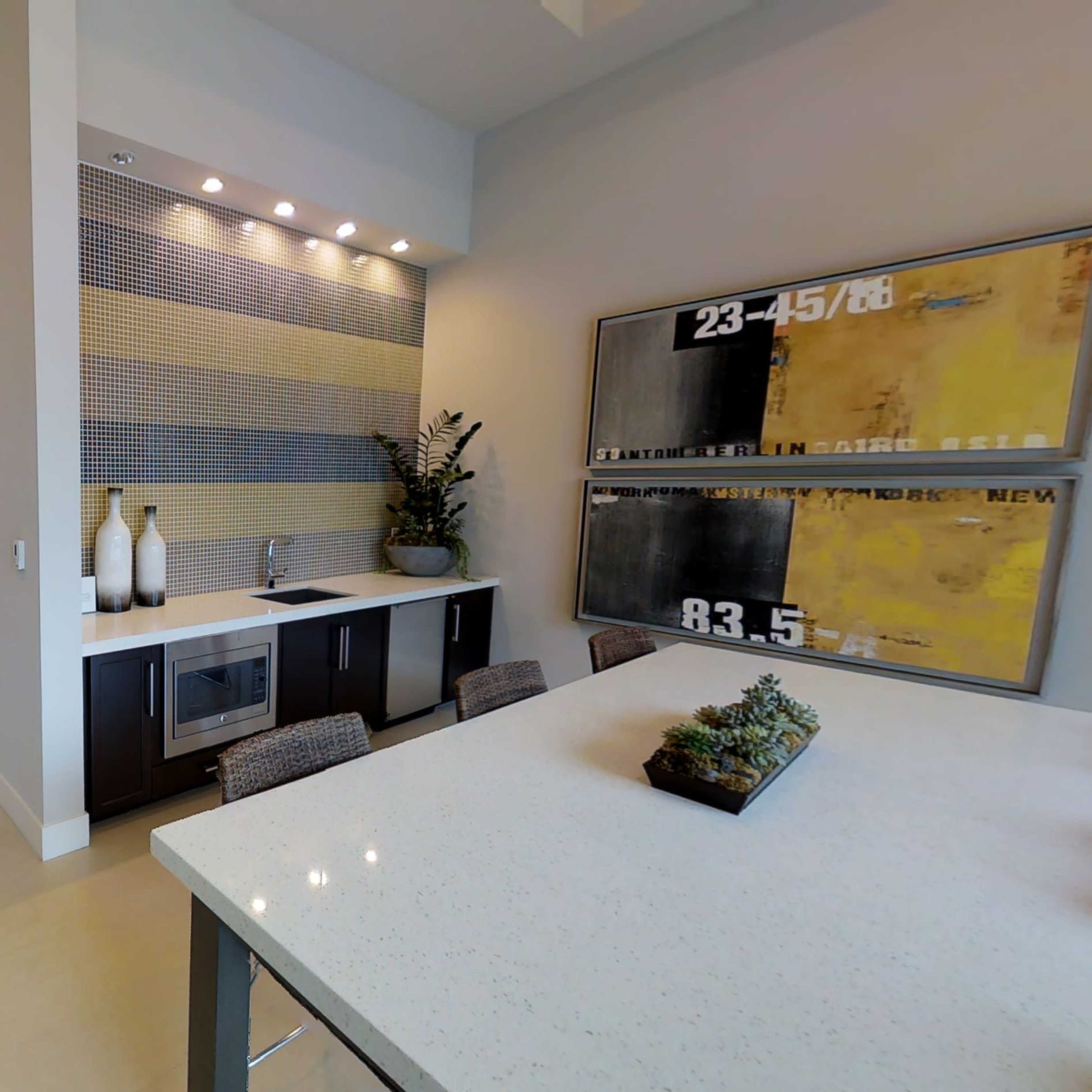 A modern interior space that includes a dining area, a kitchenette with a bar, and a living area visible in the background.