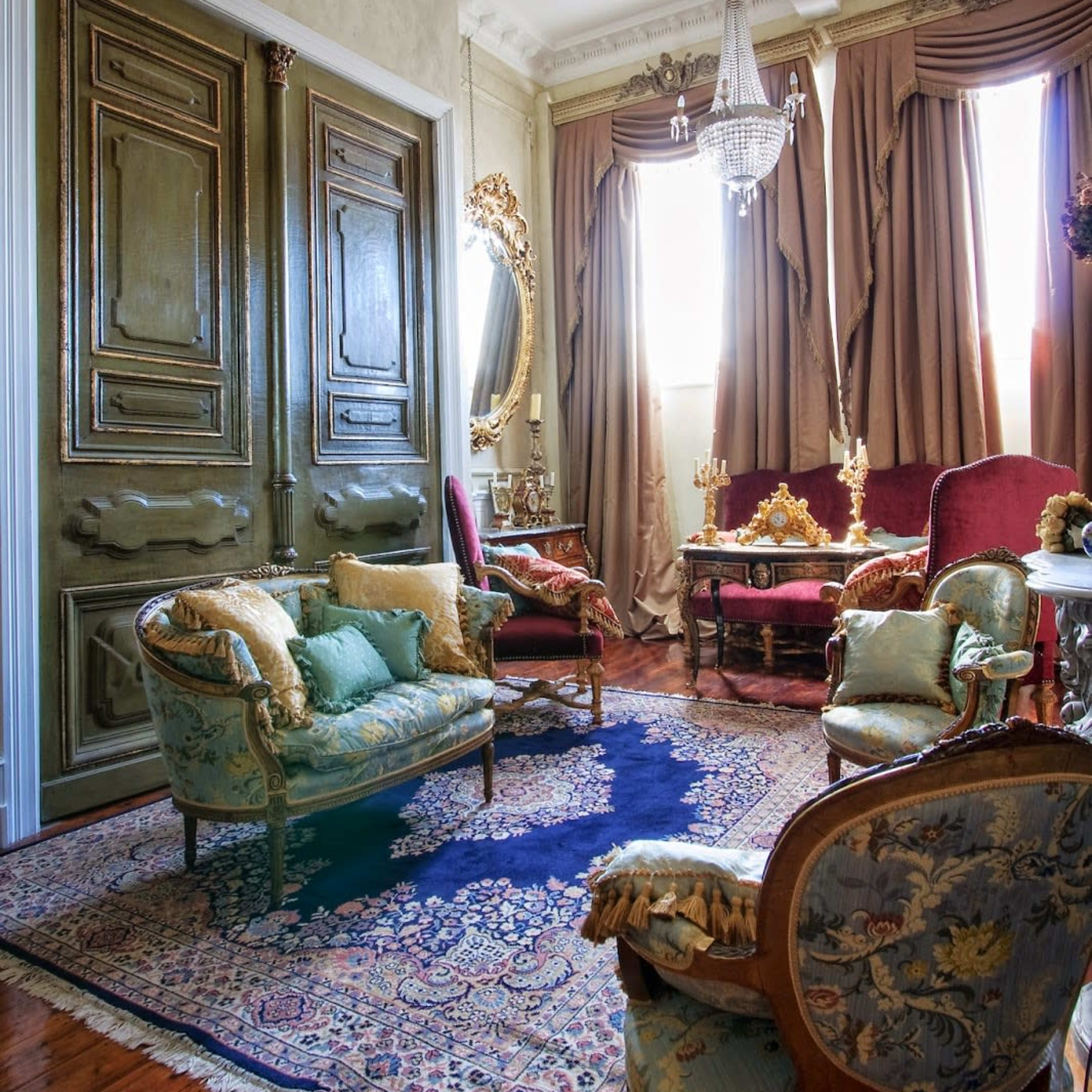 The room features ornate wooden doors, elegant seating arrangements, and large drapes with a chandelier illuminating the space.