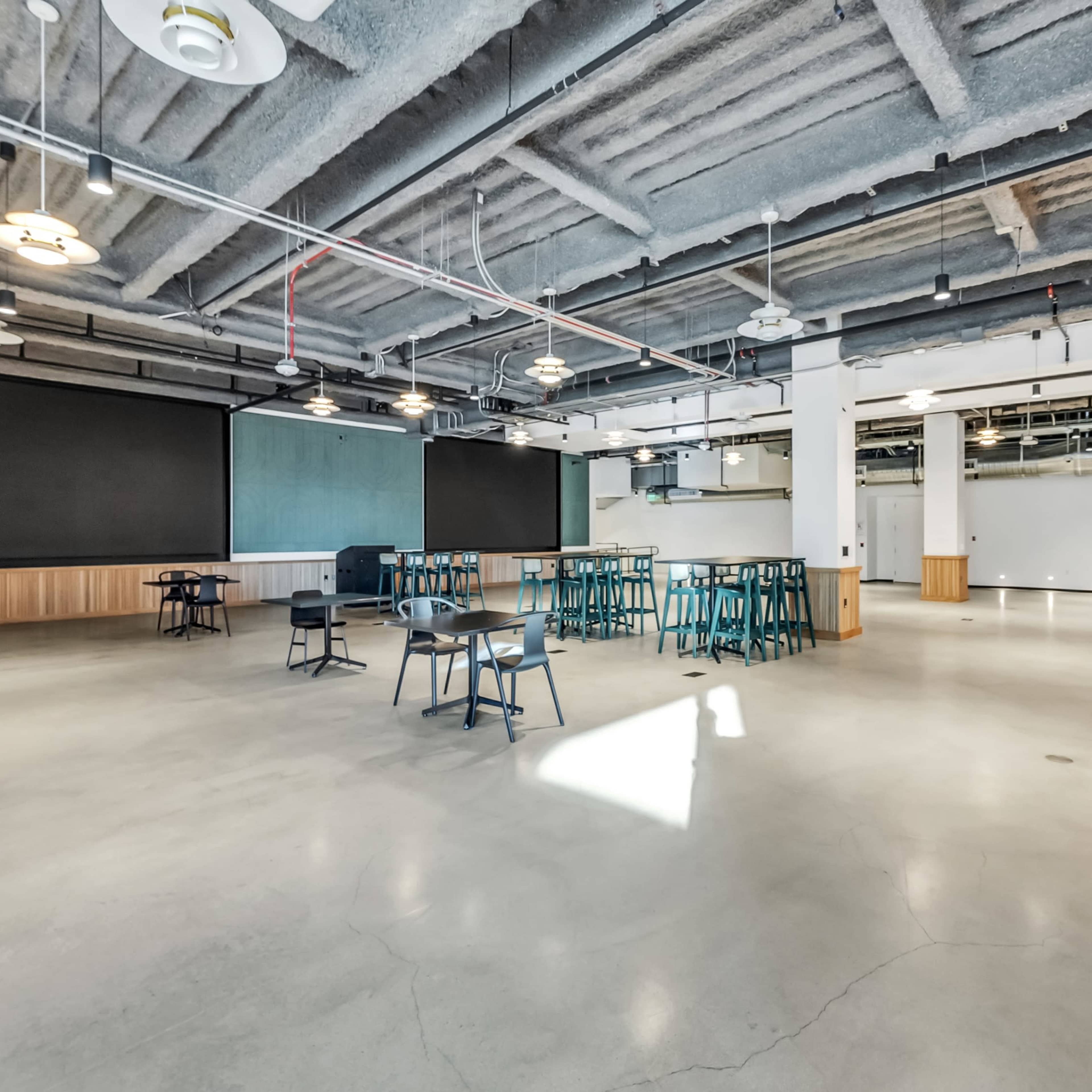 A large, open space with concrete floors, several tables and chairs, and a kitchenette area, all under a high ceiling with visible beams.