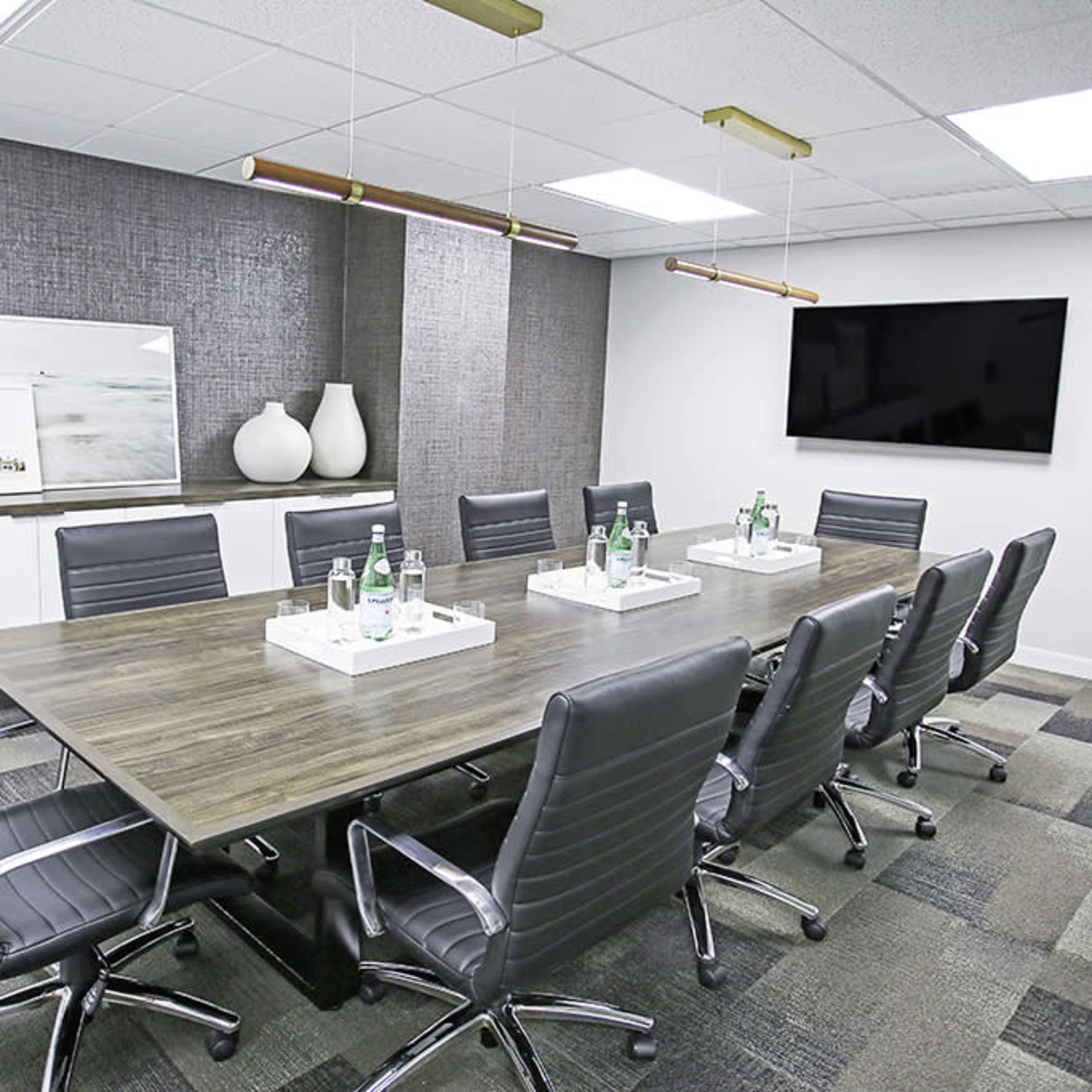 A modern conference room features a long wooden table surrounded by ergonomic chairs, with water bottles on white trays and wall-mounted artwork.