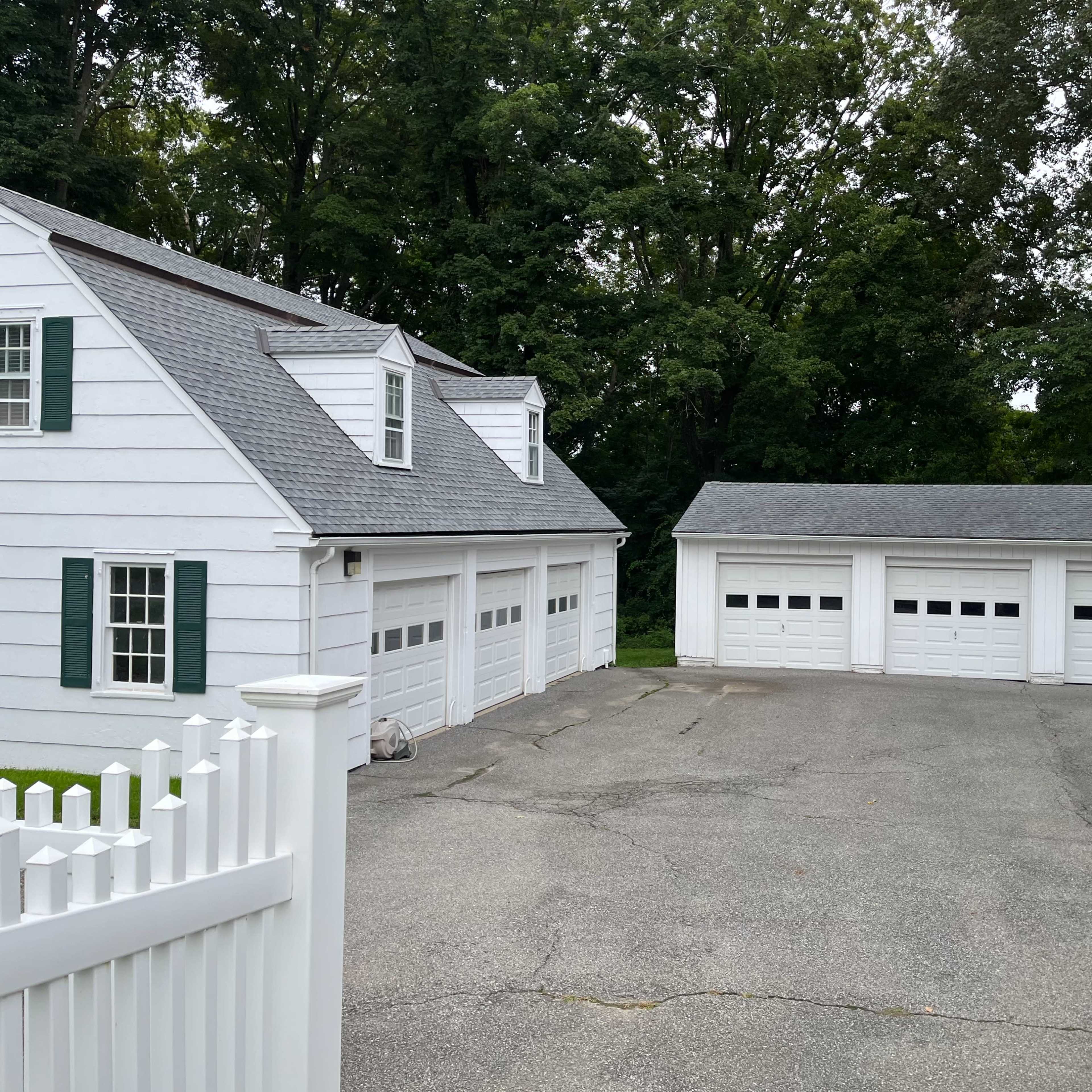 A white house with a gray roof and green shutters adjacent to a gray driveway leading to a detached garage with three closed doors.