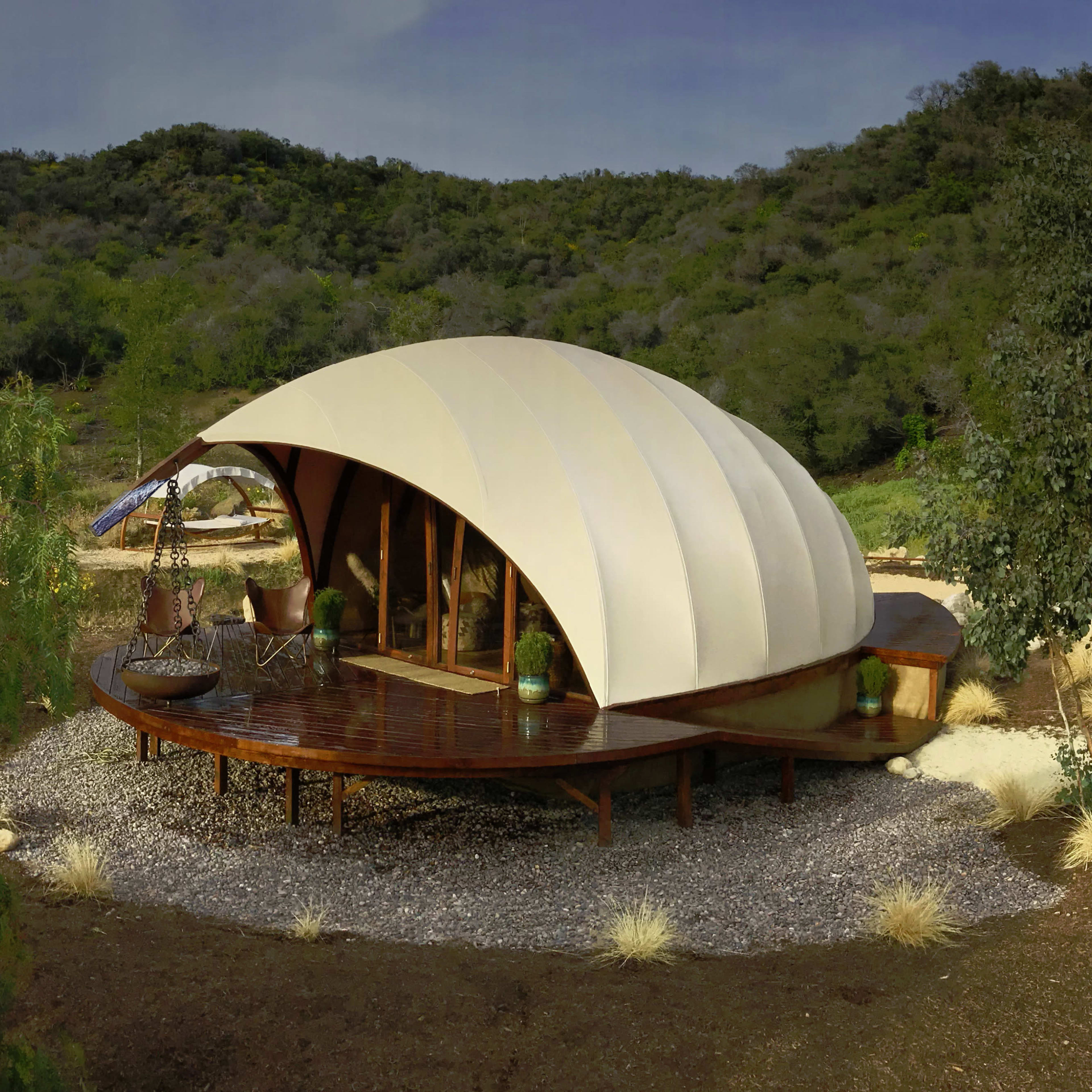 A modern, dome-shaped structure with a curved white roof situated on a wooden deck surrounded by greenery and gravel landscaping.