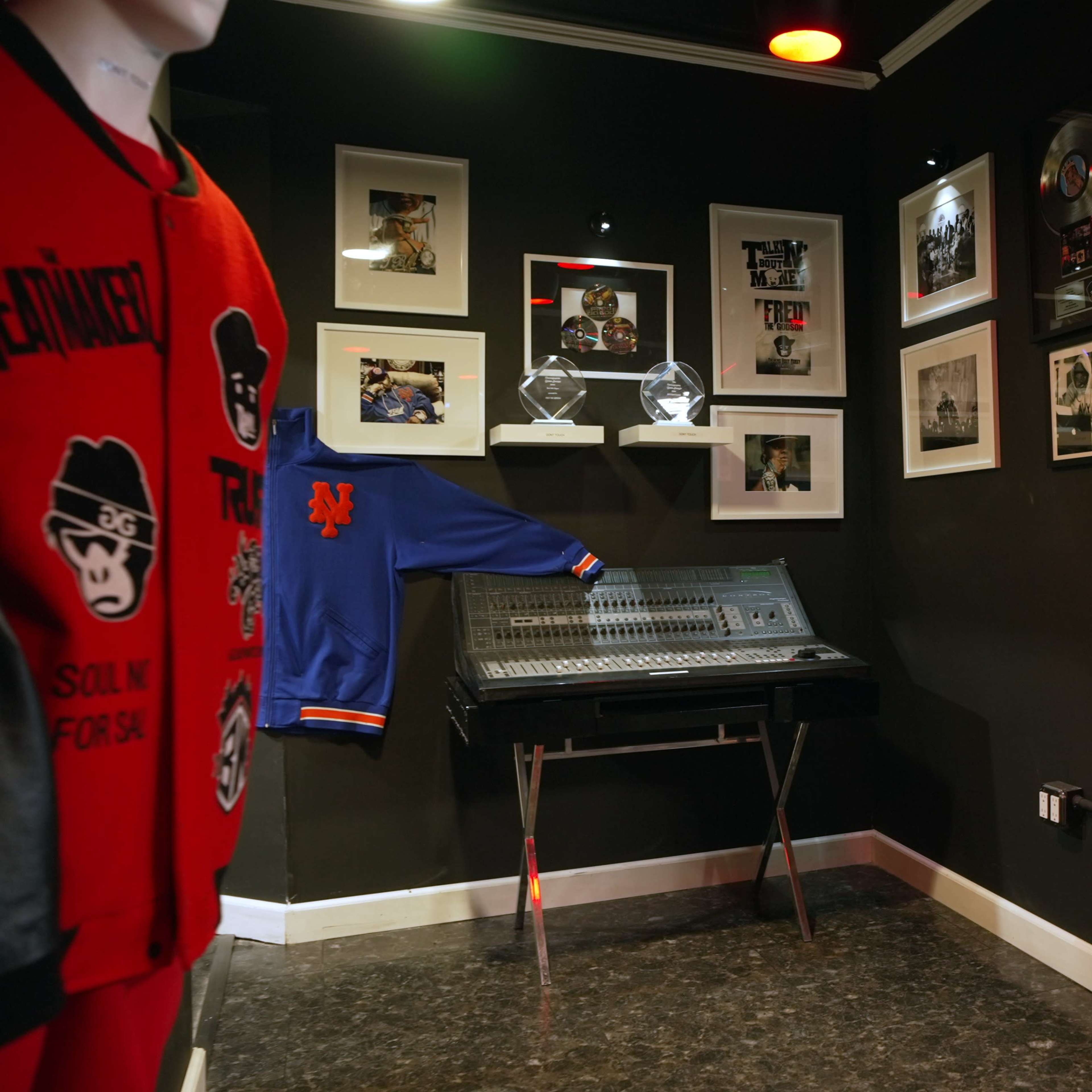 A corner of a music studio featuring a mixing console, framed photographs on the walls, and clothing displayed on a mannequin.