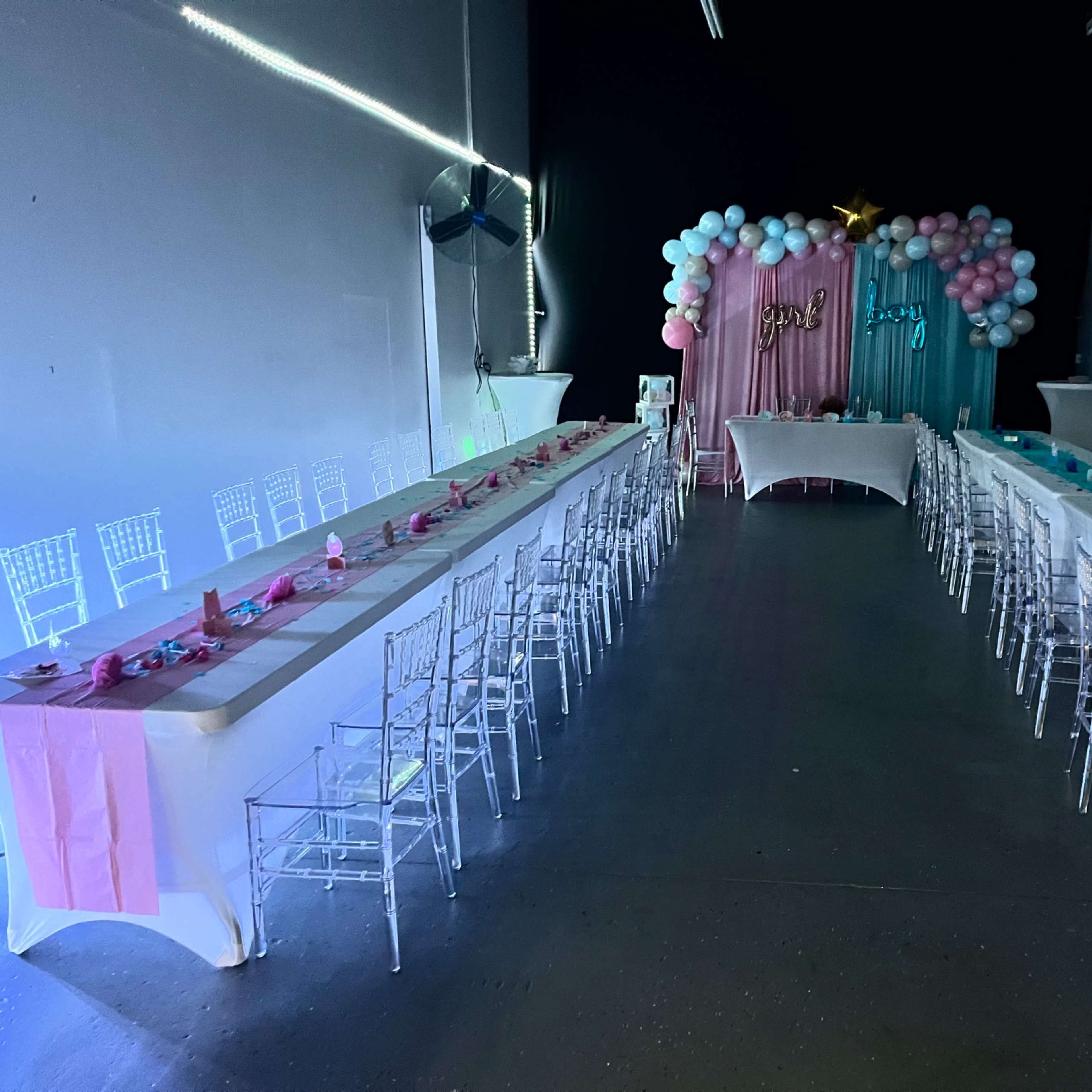 A spacious indoor venue features two long tables decorated with pink and blue accents, flanked by clear acrylic chairs, with a backdrop of balloons and a decorative stand at the front.
