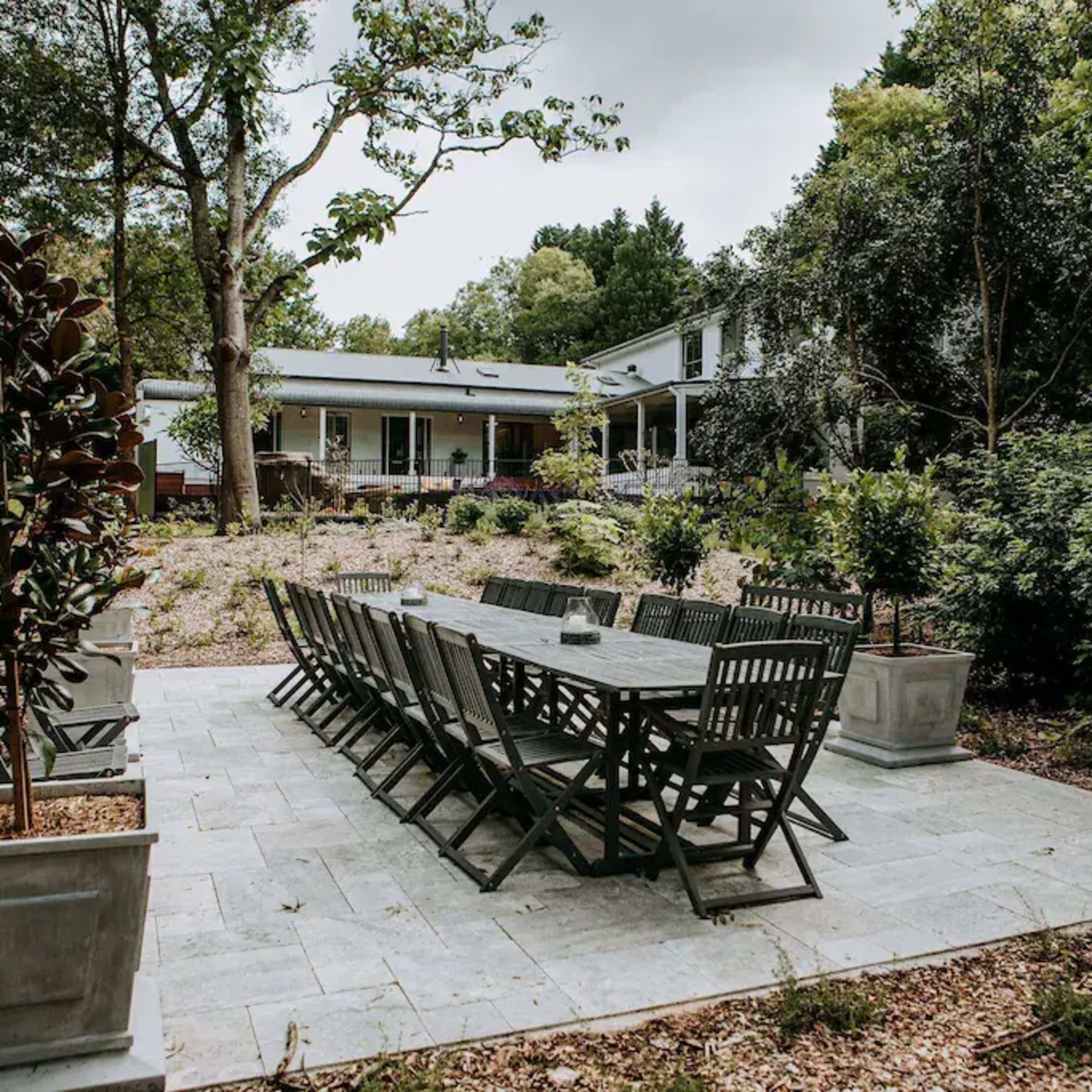A long dining table with chairs is set on a stone patio in a garden surrounded by trees and shrubs.