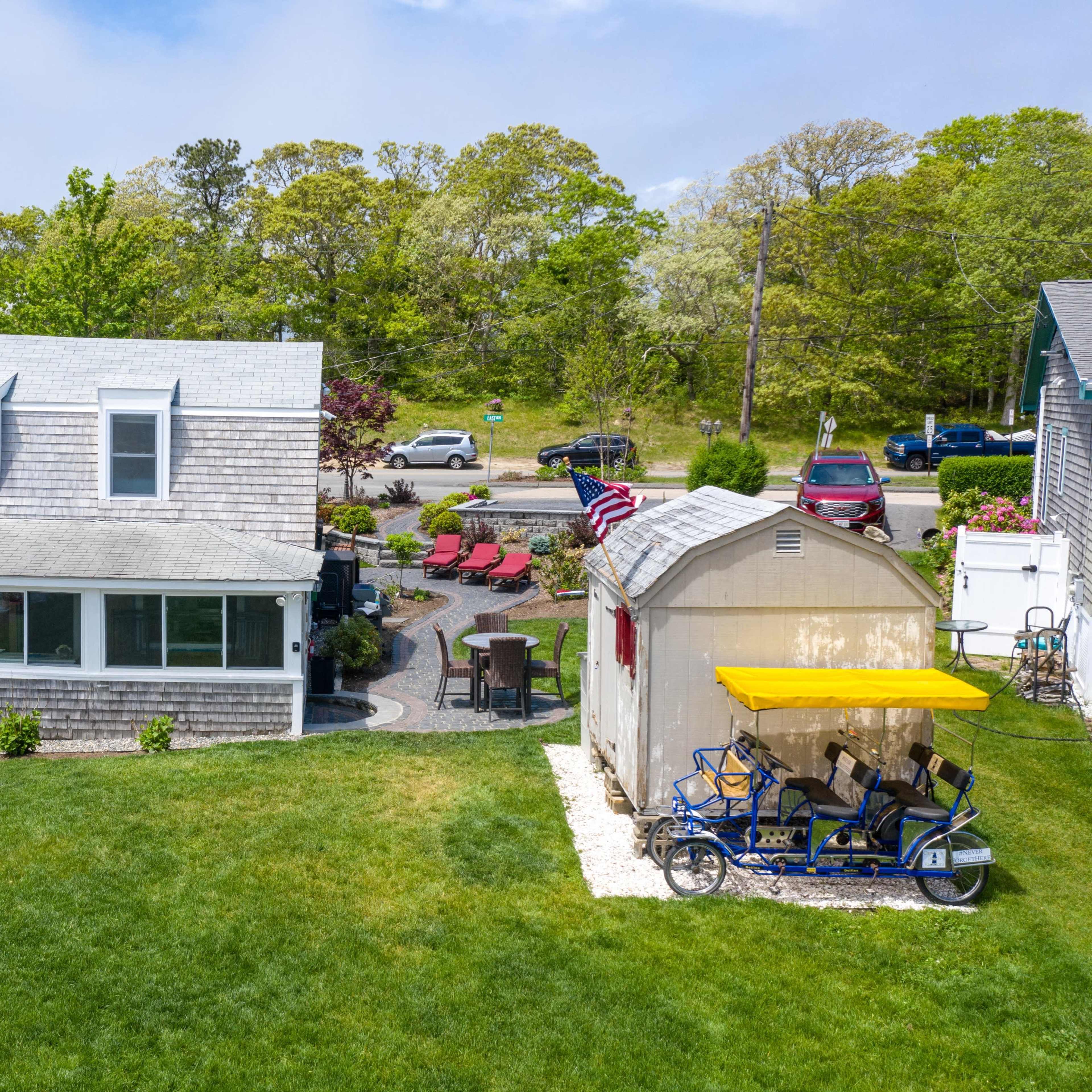 A gray-shingled house with large windows sits in a yard surrounded by green grass, patio chairs, and a storage shed with a yellow roof.