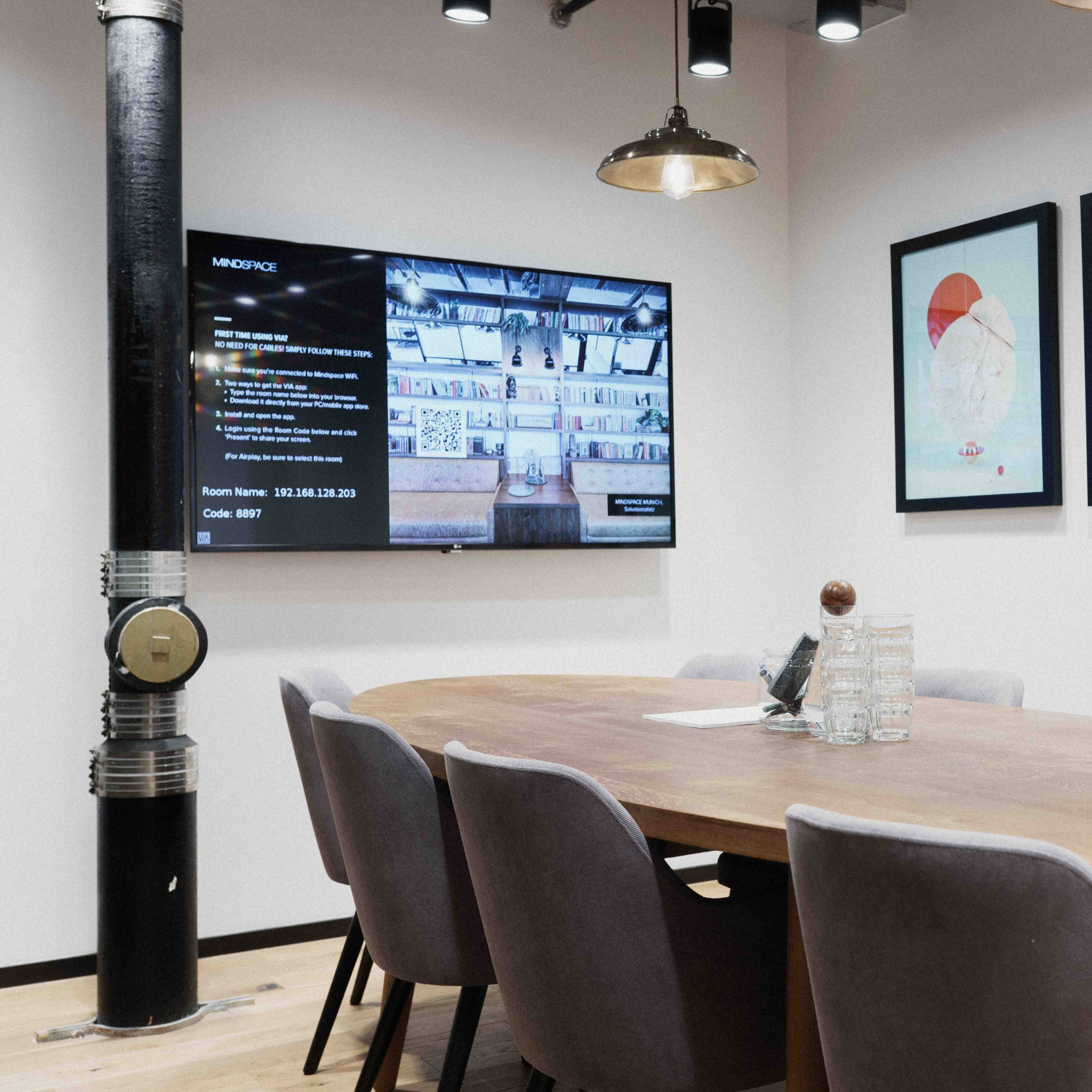 A meeting room with a large wooden table surrounded by upholstered chairs, a television displaying information on the wall, and framed artwork hanging nearby.