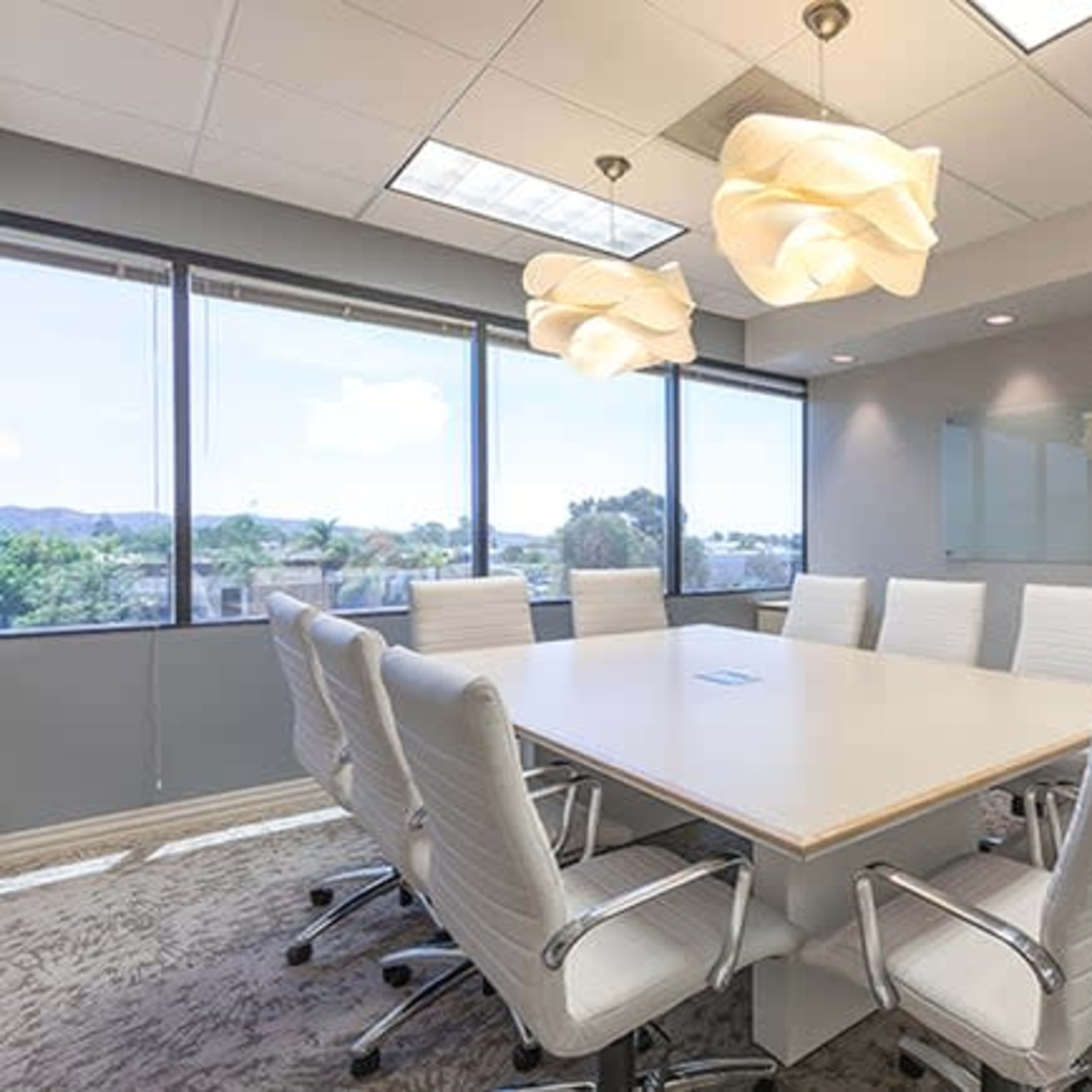 A modern conference room features a large table surrounded by white chairs, with large windows allowing natural light to enter.
