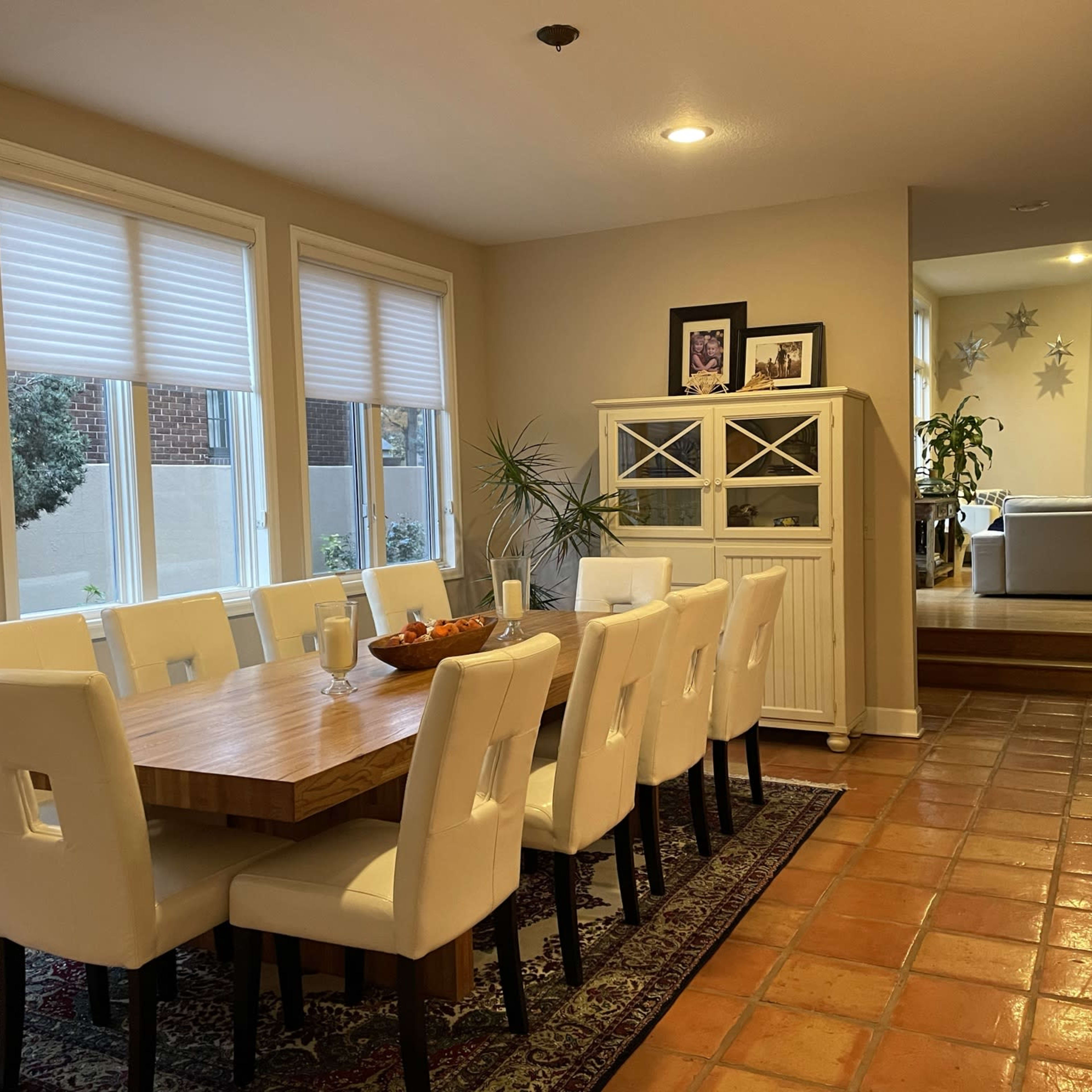 A spacious dining room features a large wooden table surrounded by white chairs, with a sideboard and windows offering views of a garden.