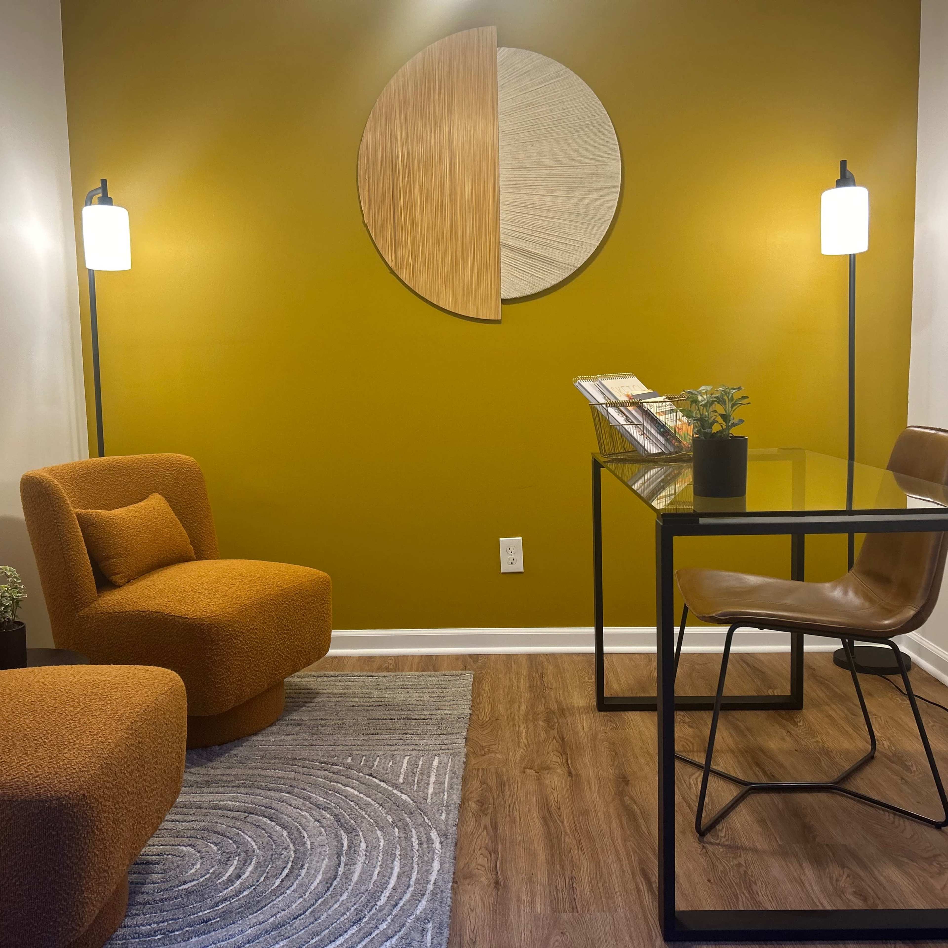A small modern office space with two orange upholstered chairs, a glass desk with a brown chair, and a decorative wall featuring a circular artwork on a mustard-colored background.