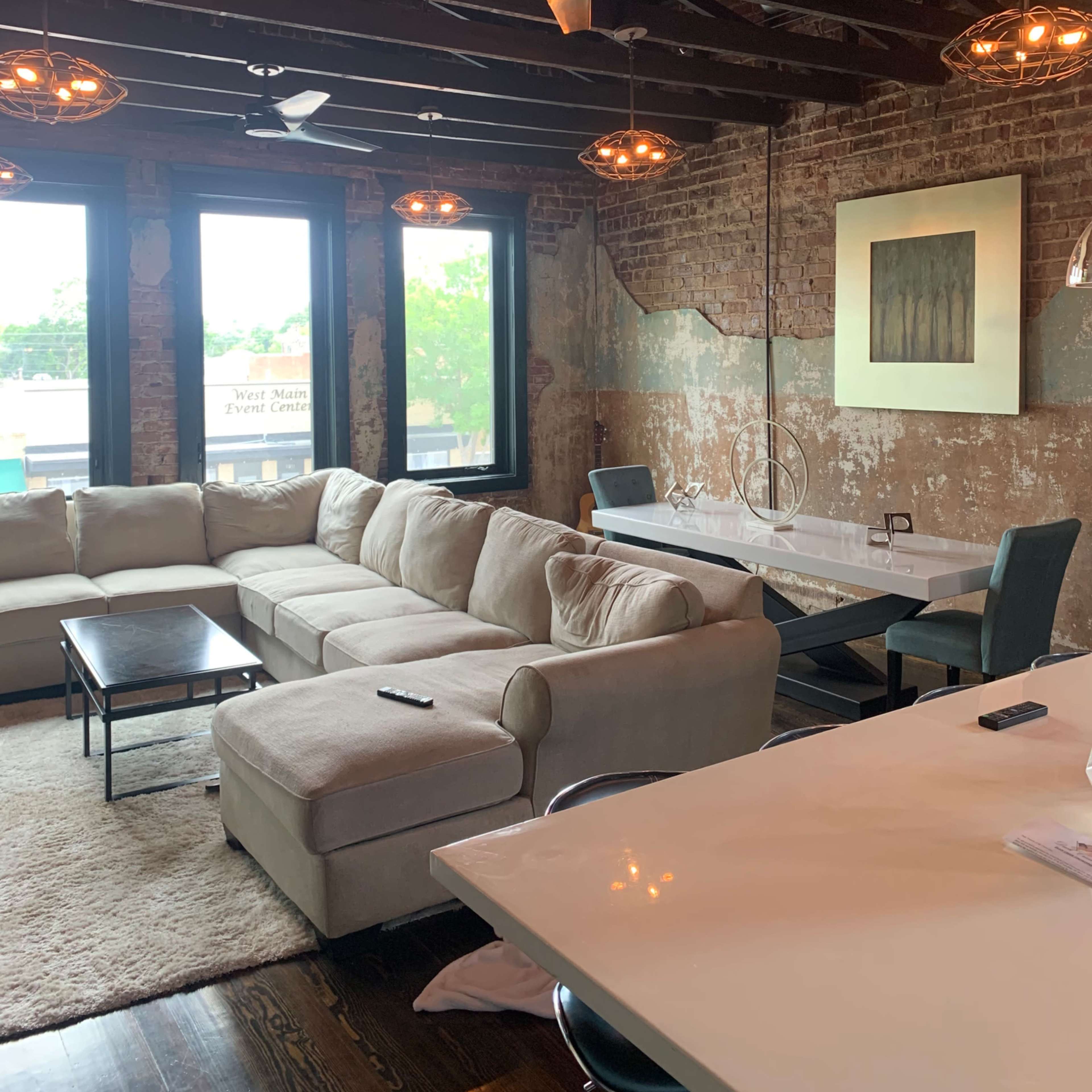 A spacious living room with exposed brick walls, large windows, a light-colored sectional sofa, and a dining table.