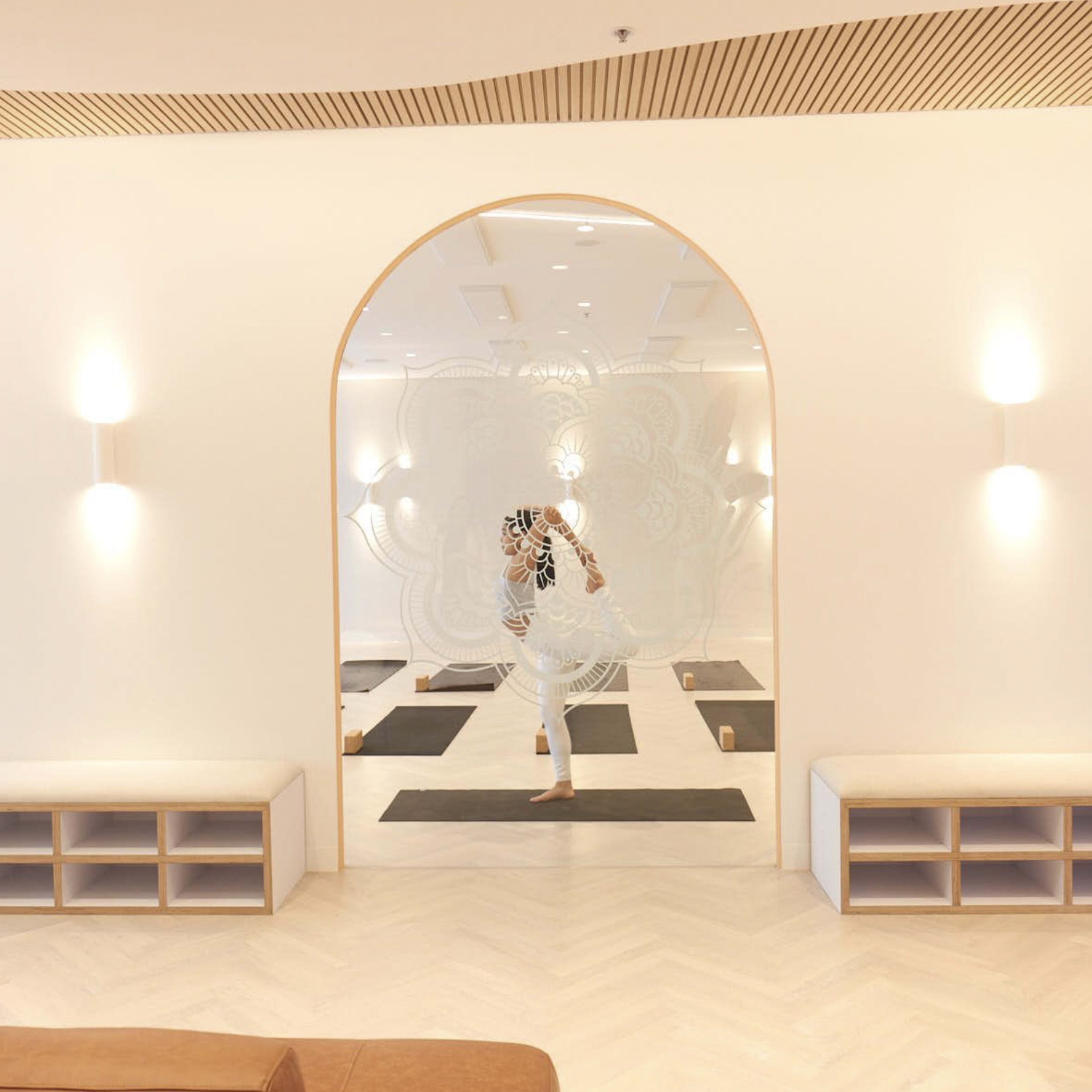 A person is practicing yoga in a minimalist studio with wooden benches and mirrored walls.