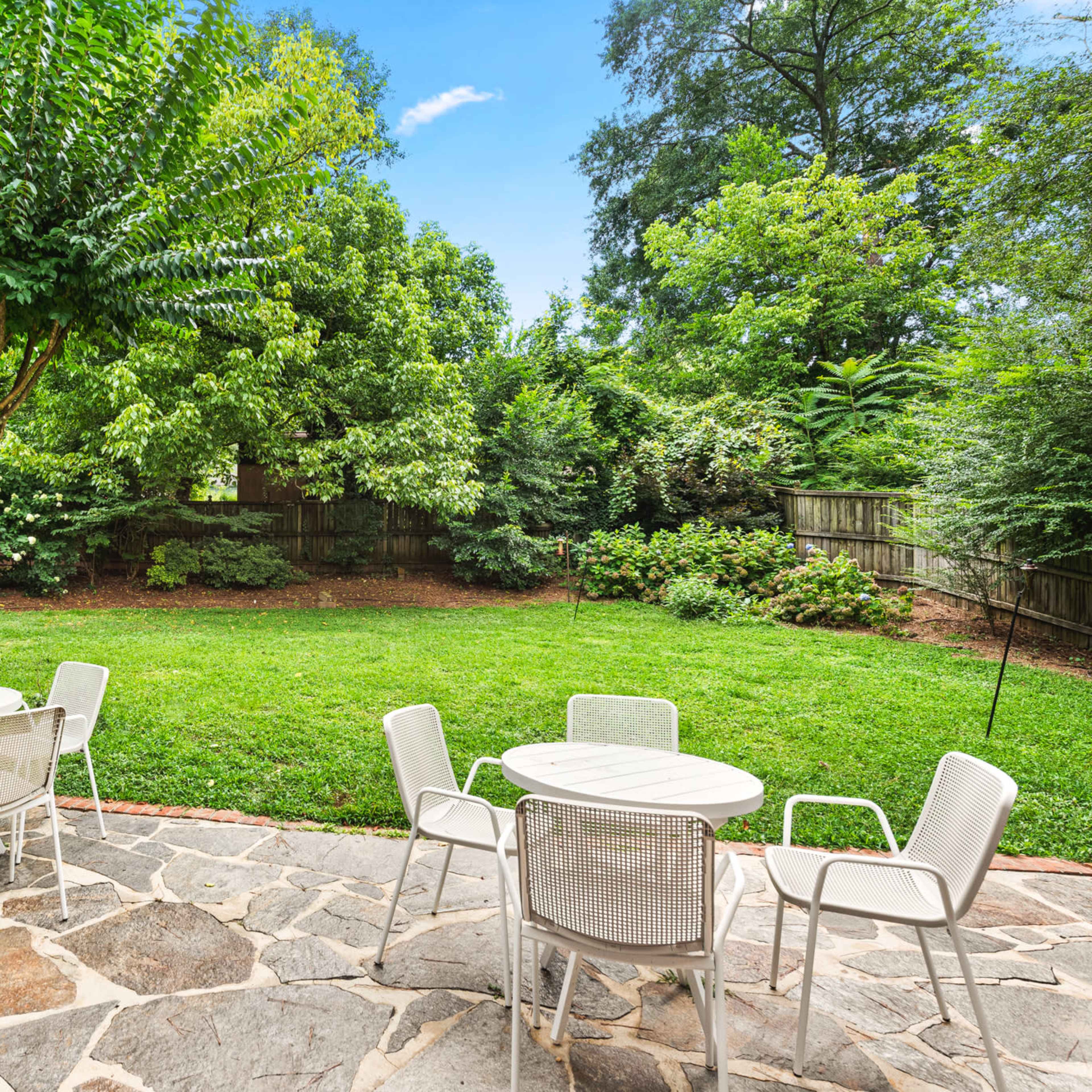 A stone patio with white outdoor furniture overlooks a grassy backyard surrounded by trees and shrubs.