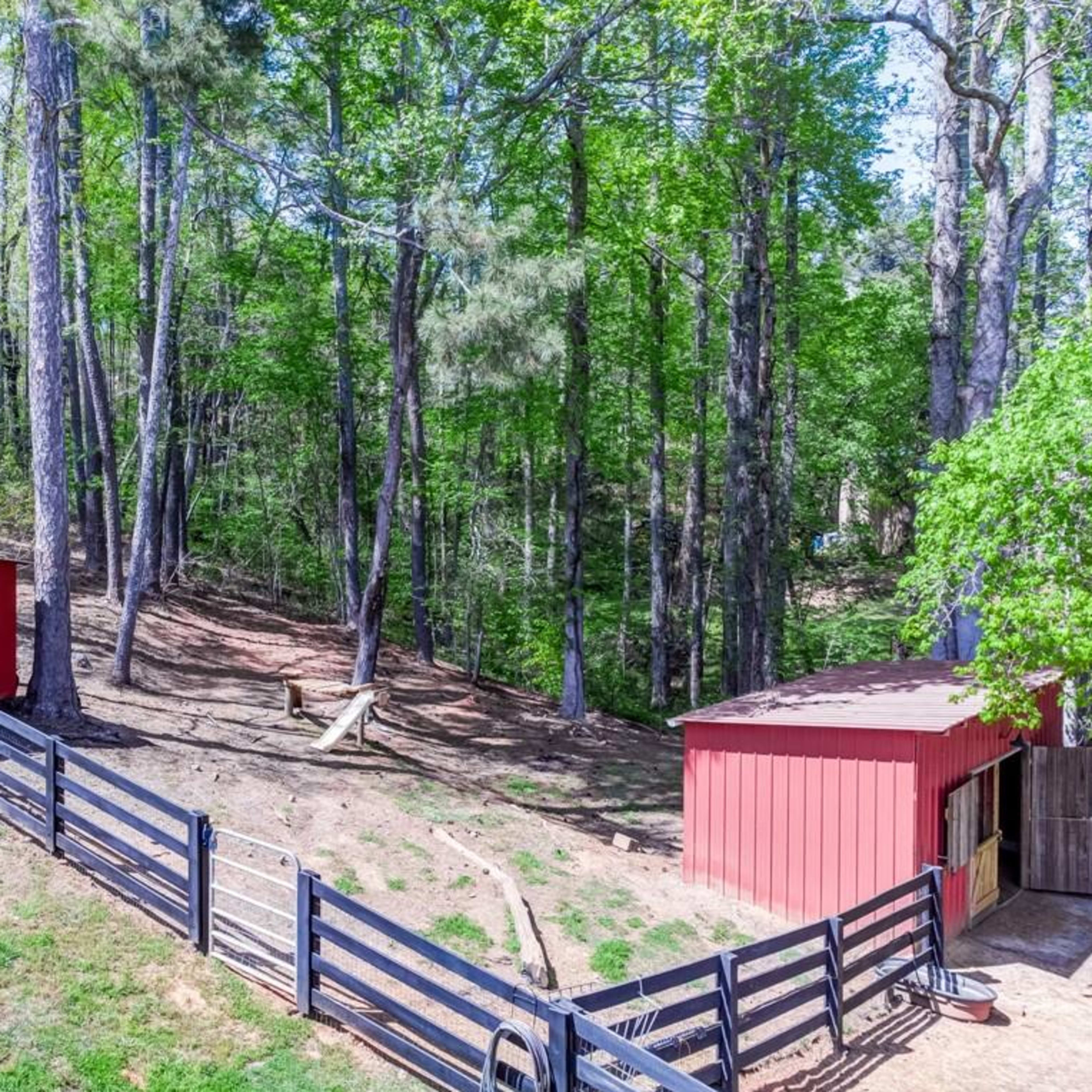 A wooded area with two red buildings and a black fence surrounding a dirt path.